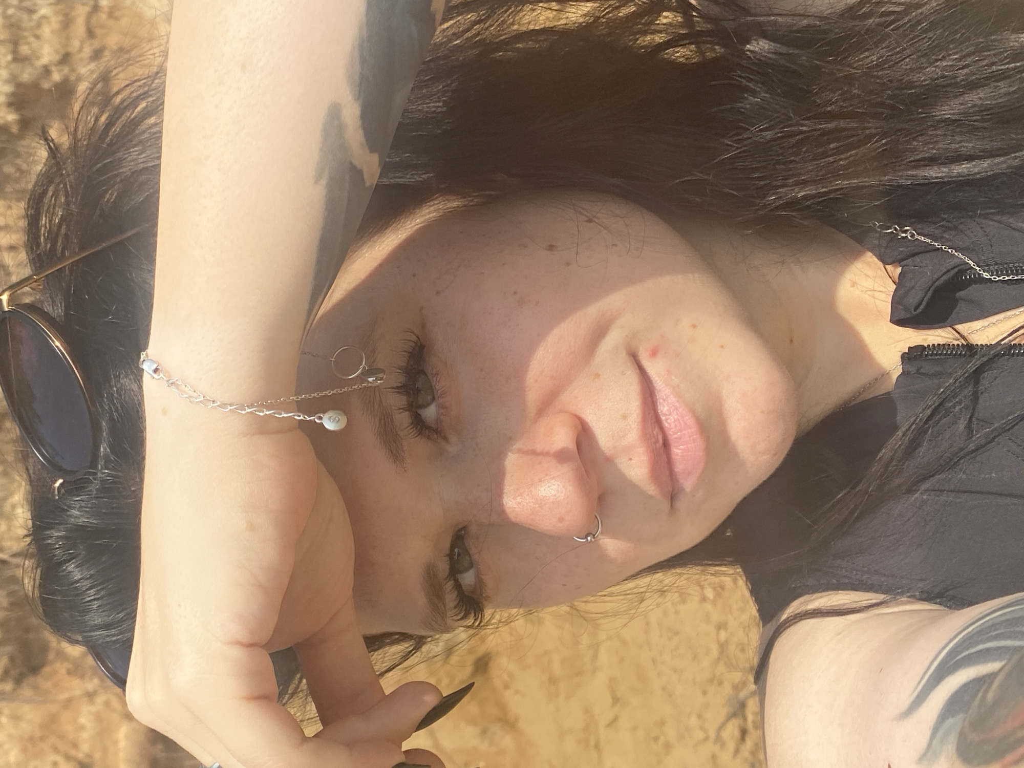 A close-up, sun-drenched selfie shows a woman with dark hair and fair skin, her left hand shielding her forehead, revealing a portion of her face. She wears sunglasses pushed back on her head, and a delicate silver bracelet adorns her wrist. Her nose is pierced with a small silver hoop, and she has prominent eyelashes. Sunlight casts shadows across her face, highlighting freckles and a single pimple on her cheek. Her attire includes a black top with a zipper. The background appears to be an outdoor environment with blurred, sandy-brown terrain, possibly a park or natural landscape. The location within Novyi, Russia is not discernible from the visual information provided.