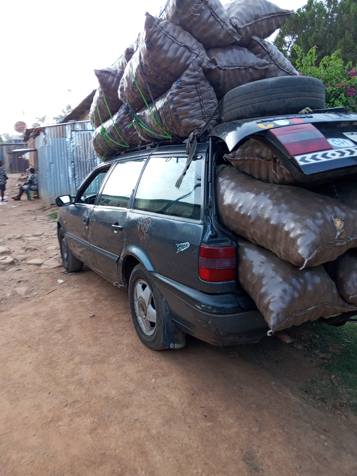 The image depicts a heavily overloaded dark blue Volkswagen Passat wagon parked on a dirt road in Bukuru, Nigeria. The car is packed with large brown sacks, stacked both on the roof and inside the rear cargo area. A spare tire rests on top of the roof cargo. The license plate is partially visible and shows "GH" and chevron patterns. The car appears old and worn. In the background, there are corrugated metal buildings and people sitting on a bench. The environment looks like a rural or less developed area.