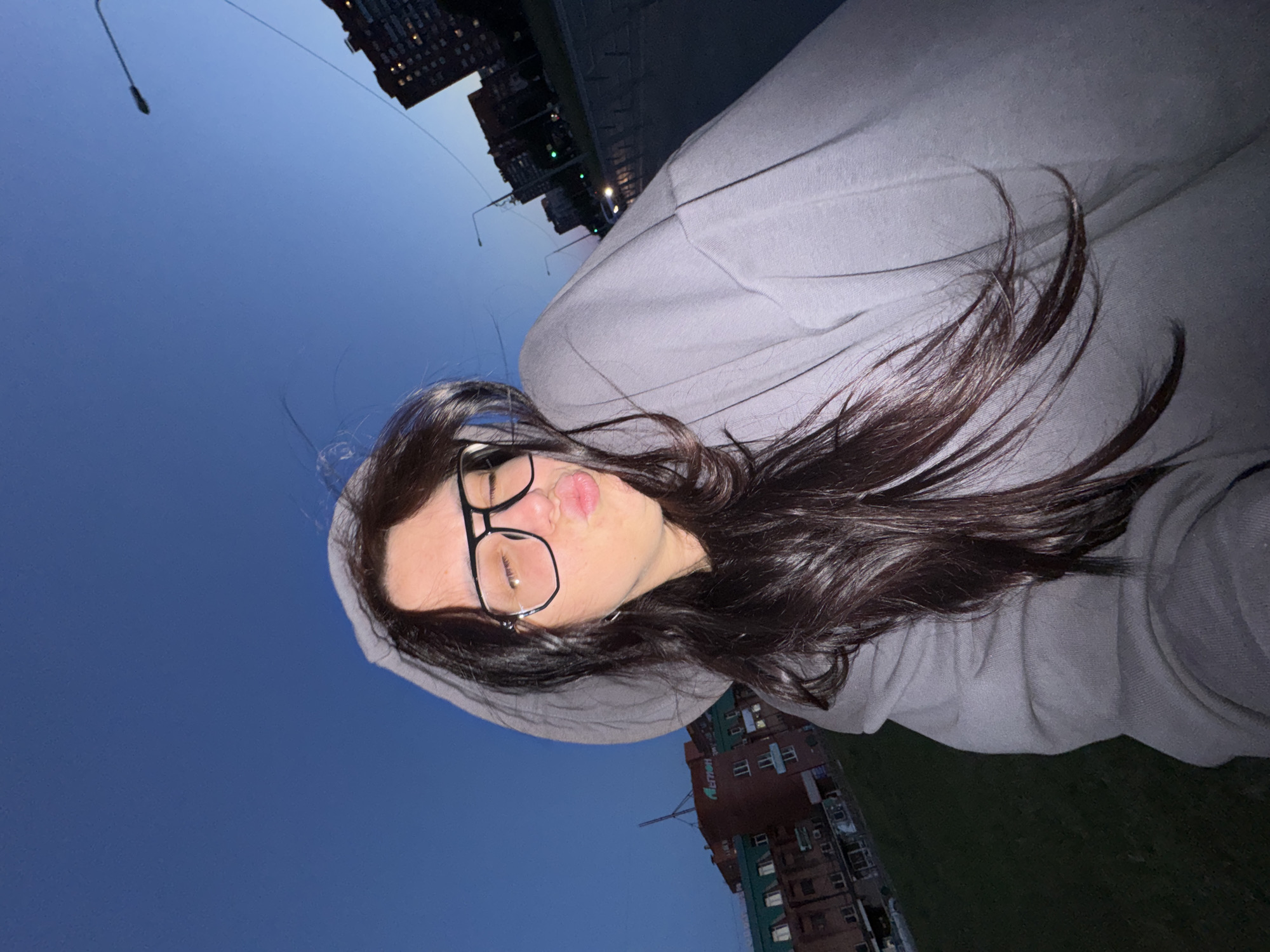 A young woman is captured in a close-up selfie taken outdoors during dusk or twilight. The camera is held at a slight upward angle, showing her from the shoulders up, with her long, dark brown hair flowing around her face. She is wearing clear-rimmed glasses with dark frames and a light grey hooded sweatshirt, with the hood pulled up around her head. Her lips are playfully pursed in a 'duck face' pose, and she is looking directly at the camera.
The background features a deep blue sky, indicating the transition from day to night. Several multi-story urban buildings, some with illuminated windows, are visible against the skyline. Faint streetlights can also be seen in the distance. Below the buildings, there is a strip of dark ground or grass, suggesting a park or open area. Notably, on one of the buildings in the background, the Cyrillic text "МЕЧТАЙ" (MECHTAY), meaning "DREAM" in Russian, is faintly visible. The overall impression is a casual, self-portrait taken in an urban environment as evening sets in.
