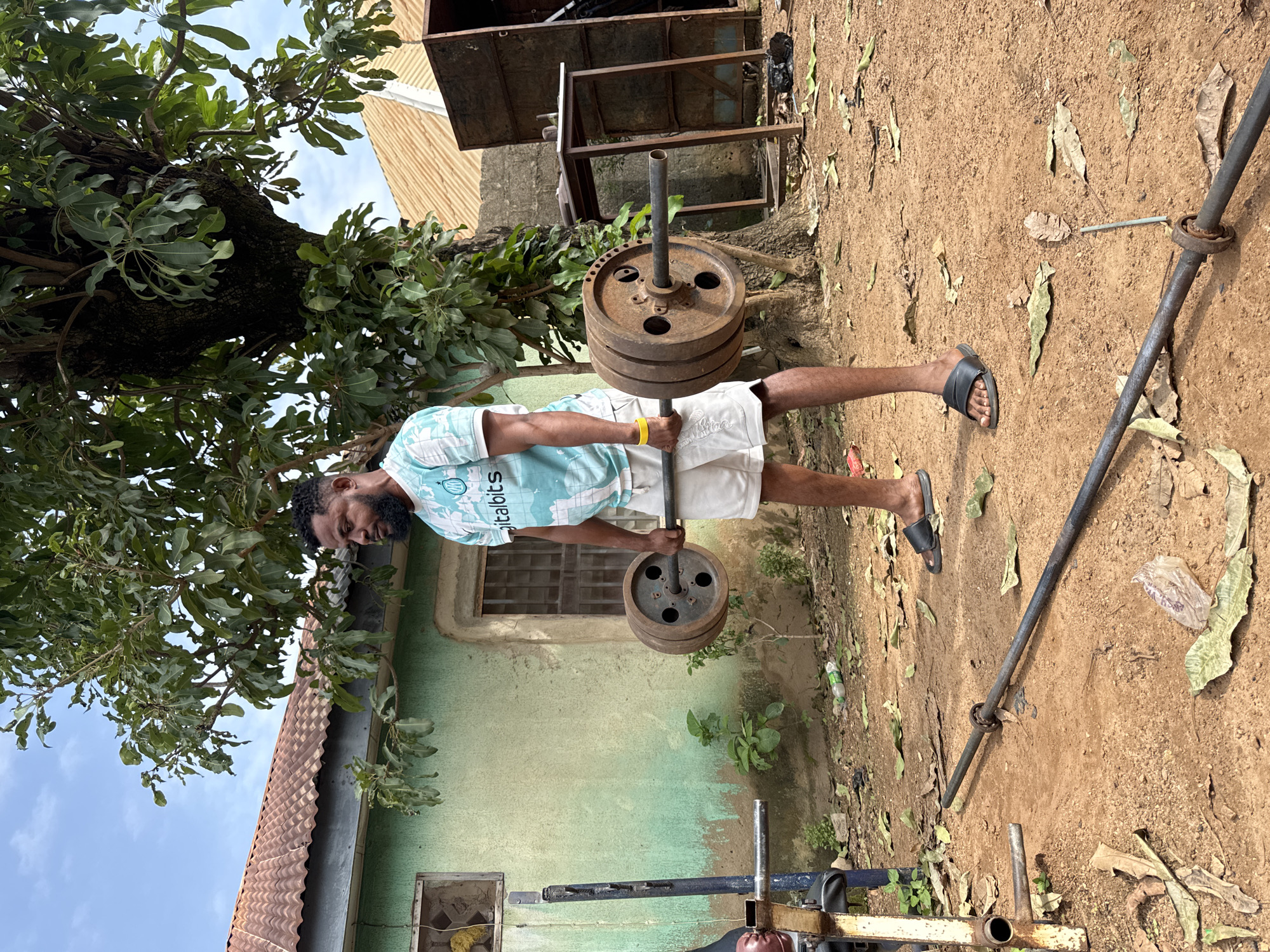 A man stands outdoors in what appears to be a backyard in Akwanga, Nigeria, performing a deadlift with a barbell. He is wearing a light-colored t-shirt with a blue and white pattern and white shorts, along with black sandals. A yellow band is visible on his left wrist. The barbell is loaded with heavy, circular weight plates. The environment is rustic, with a light green painted building behind him, a large tree to his right, and dry leaves scattered on the dirt ground. Other objects in the scene include weightlifting equipment, a metal table, and some debris. The sky is partly cloudy.