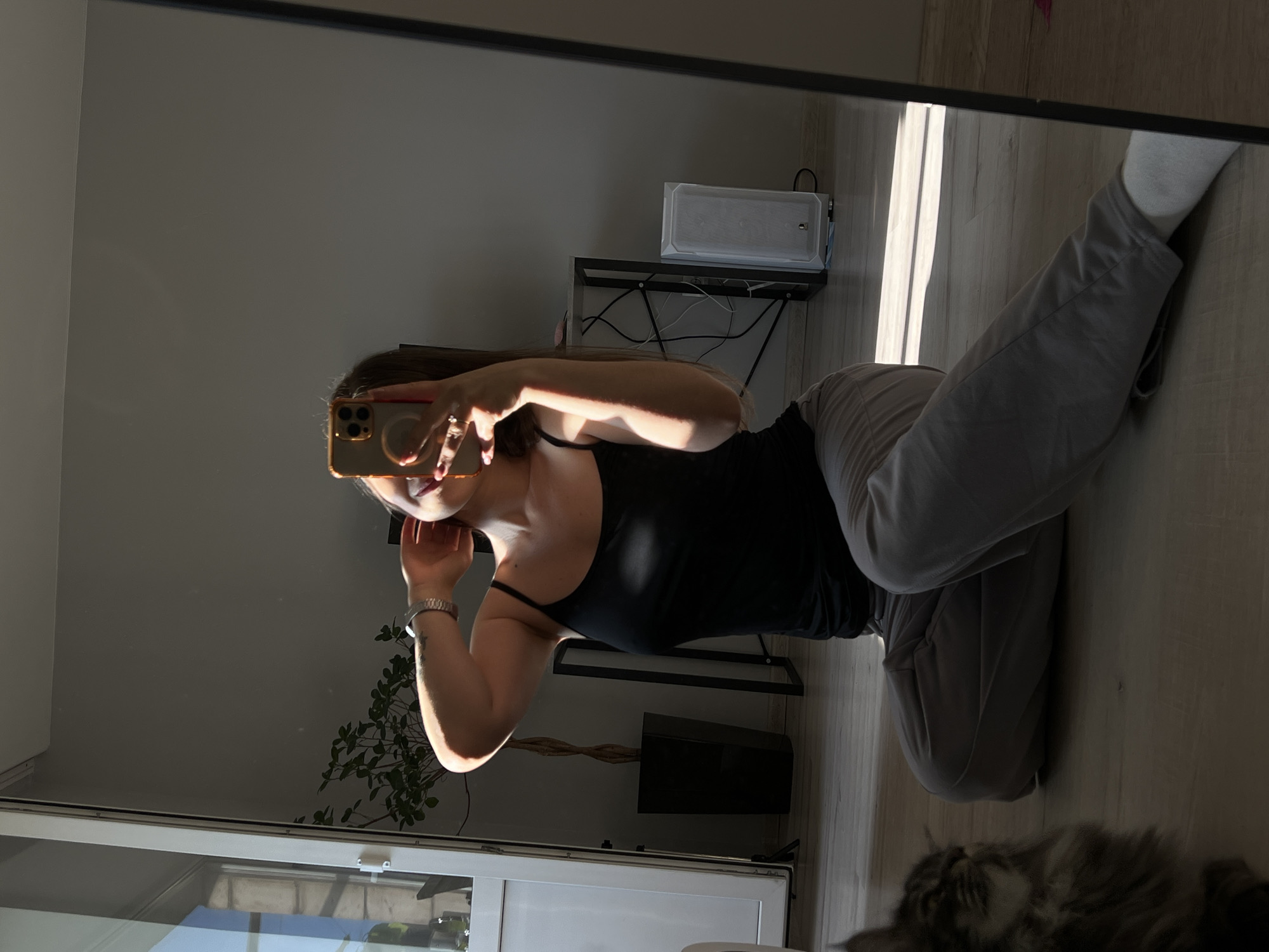 A woman is taking a mirror selfie while sitting cross-legged on a wooden floor. She is wearing a black tank top and grey sweatpants, with white socks. The woman is holding her phone in her right hand, and her left hand is behind her neck. Sunlight is streaming in from the left, casting shadows on the floor and wall. In the background, there is a potted plant, a black shelving unit with a computer tower, and a white speaker. A cat is partially visible in the lower left corner of the image. The setting appears to be an indoor residential space. There is no discernible indication of the location being in Ternopil, Ukraine.