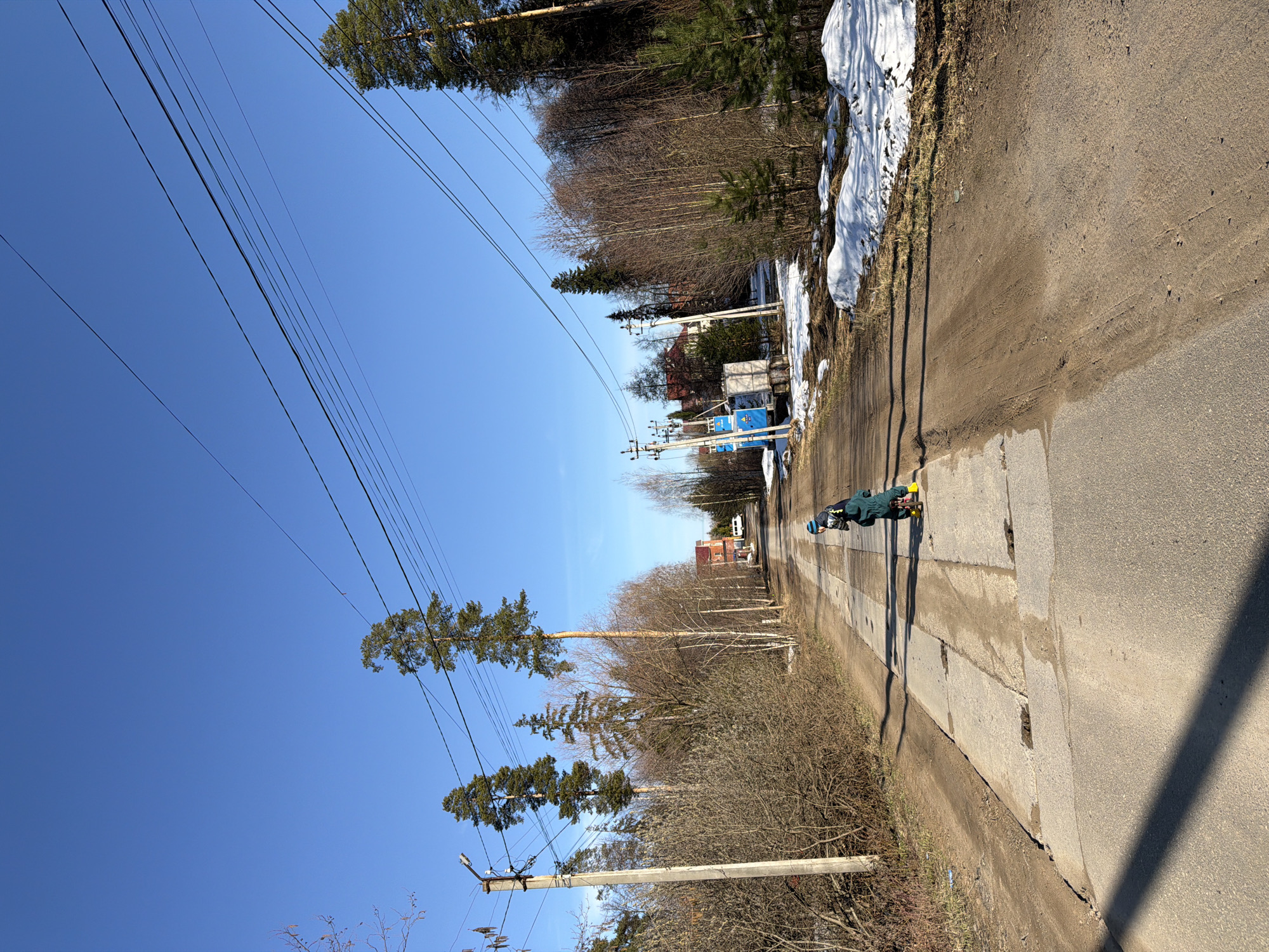 The image shows a street in Chaikovsky, Russia, on a sunny day. A person wearing a green jacket and yellow shoes is standing on the sidewalk along the road. Patches of snow are visible on the right side of the road. Overhead power lines stretch across the clear blue sky. Bare trees and some tall pines line the road. A concrete curb is visible alongside the asphalt.