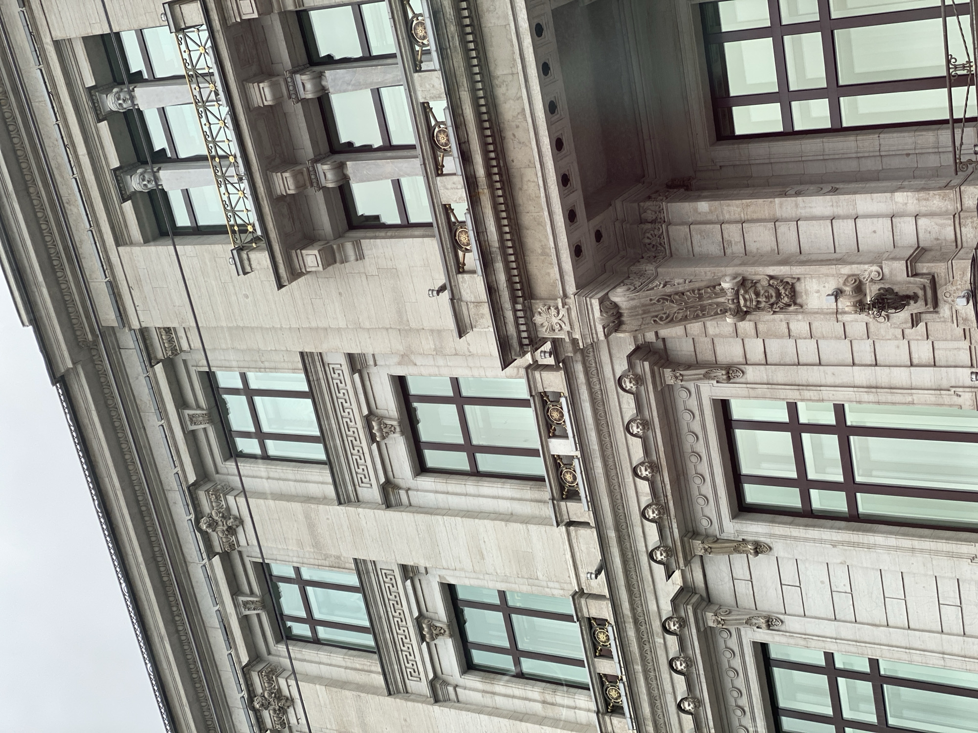 A low-angle, close-up shot reveals the architectural details of a building facade in an urban setting. The building, likely located in an area with significant historical construction, is characterized by its ornate stone detailing, including decorative moldings, repetitive geometric patterns, and carved embellishments between window openings. The windows are modern, with dark frames contrasting against the light-colored stone. The sky is overcast. A metal framework attached to the building suggests a historical or decorative elevator.