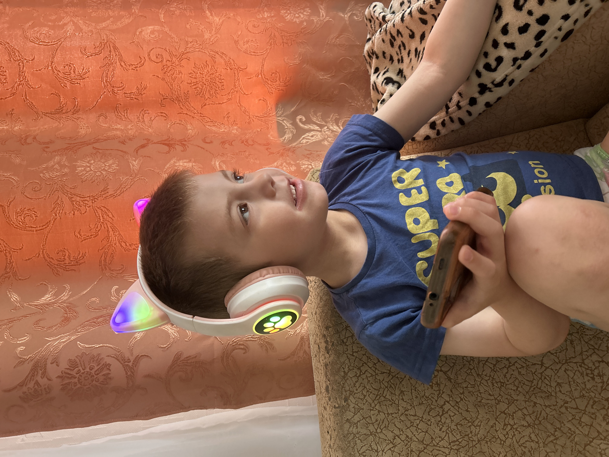 A young boy wearing a blue t-shirt and headphones with illuminated cat ears is lying on a couch, holding a mobile phone. The couch has a patterned upholstery in beige and brown tones. An orange patterned wallpaper is visible in the background. Part of a leopard print blanket is also visible in the upper right corner. The boy is smiling and looking upwards. The location is Фоки, Russia.
