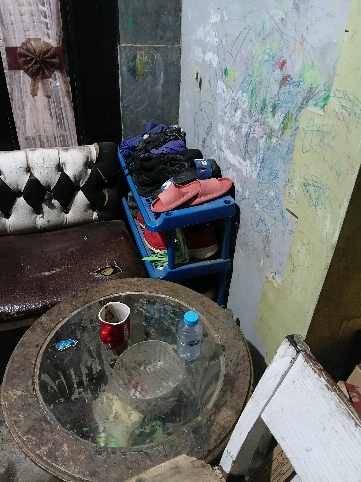 The image depicts an indoor scene, likely a residential setting in Cikal, Indonesia. In the foreground, a circular wooden table with a glass top is visible. On the table, there is a red mug containing a dark substance, possibly coffee grounds, a small blue-capped bottle of water, and a foil wrapper from a candy or snack. To the left of the table, a portion of a sofa is visible. The sofa has a brown leather seat with a visible tear and a tufted back with white and black upholstery. Behind the sofa, a blue plastic shelving unit holds various items of footwear, including black sneakers and pink slides. The wall behind the shelving unit is covered in colorful crayon scribbles. To the left of the sofa, a sheer white curtain with a decorative burgundy bow is hanging. The overall environment appears somewhat cluttered and unkempt, suggesting a lived-in space.