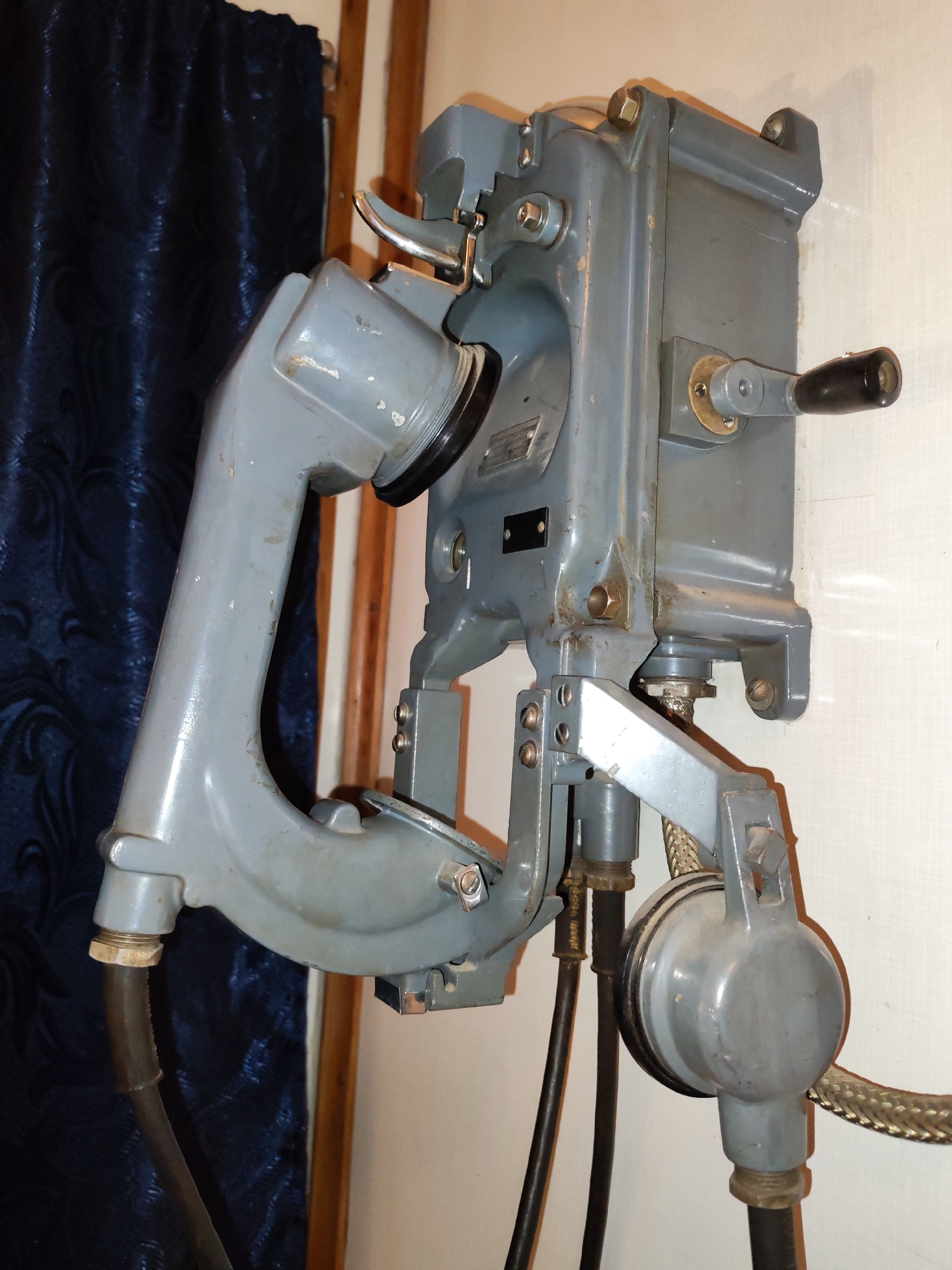 A gray, vintage, wall-mounted telephone is mounted to a light beige wall. The phone has a large earpiece with a black edge. A black cable extends from the bottom of the earpiece. The phone is secured by a gray metal bracket. A crank with a black handle is attached to the side of the phone's body. Additional black cables are connected to the bottom of the phone. A dark blue curtain hangs behind the telephone. Wooden trim is visible behind the curtain. The photo was taken indoors. There is no evidence suggesting this photograph was taken in Bol'shie Mkhi, Russia.