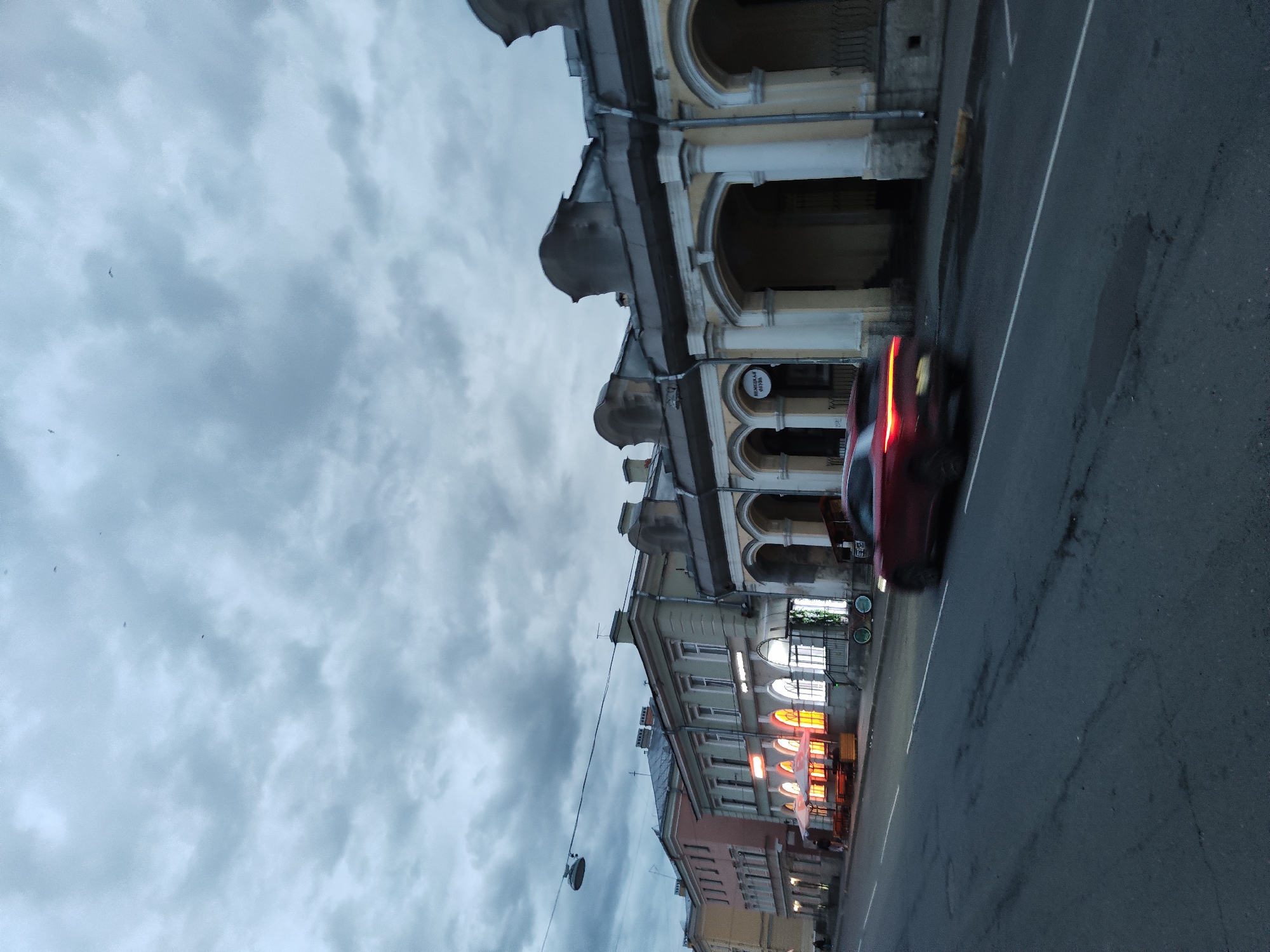 A low-angle shot depicts a street scene in Pushkin, Russia, under a cloudy sky. Architectural details reveal ornate buildings with arched facades, columns, and decorative molding lining the street. Streetlights and illuminated windows of businesses cast a warm glow against the dim, overcast backdrop. The presence of a moving car with its taillights blurred suggests active street traffic.