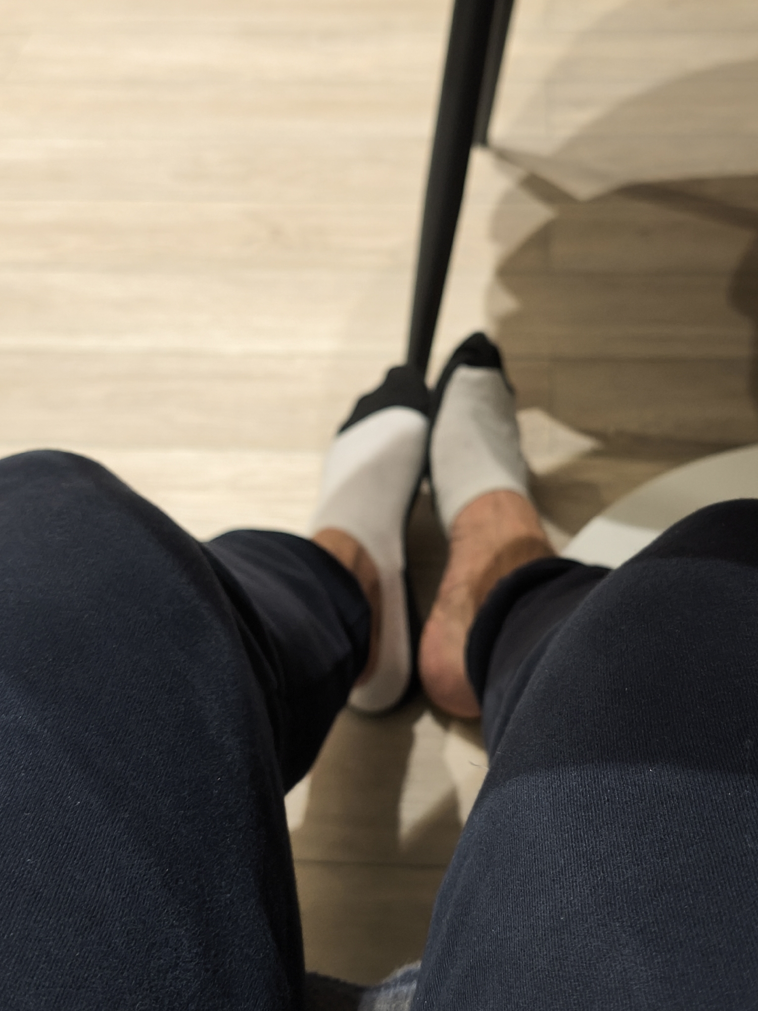 A first-person perspective photograph shows a person sitting down with their legs outstretched. They are wearing navy blue sweatpants. Their feet are in mismatched socks; one is white with a black toe, and the other is gray with a black toe. The floor is light-colored wood laminate. Part of a black chair leg is visible. The scene is likely indoors. There is no information to ascertain the location in Moscow, Russia.