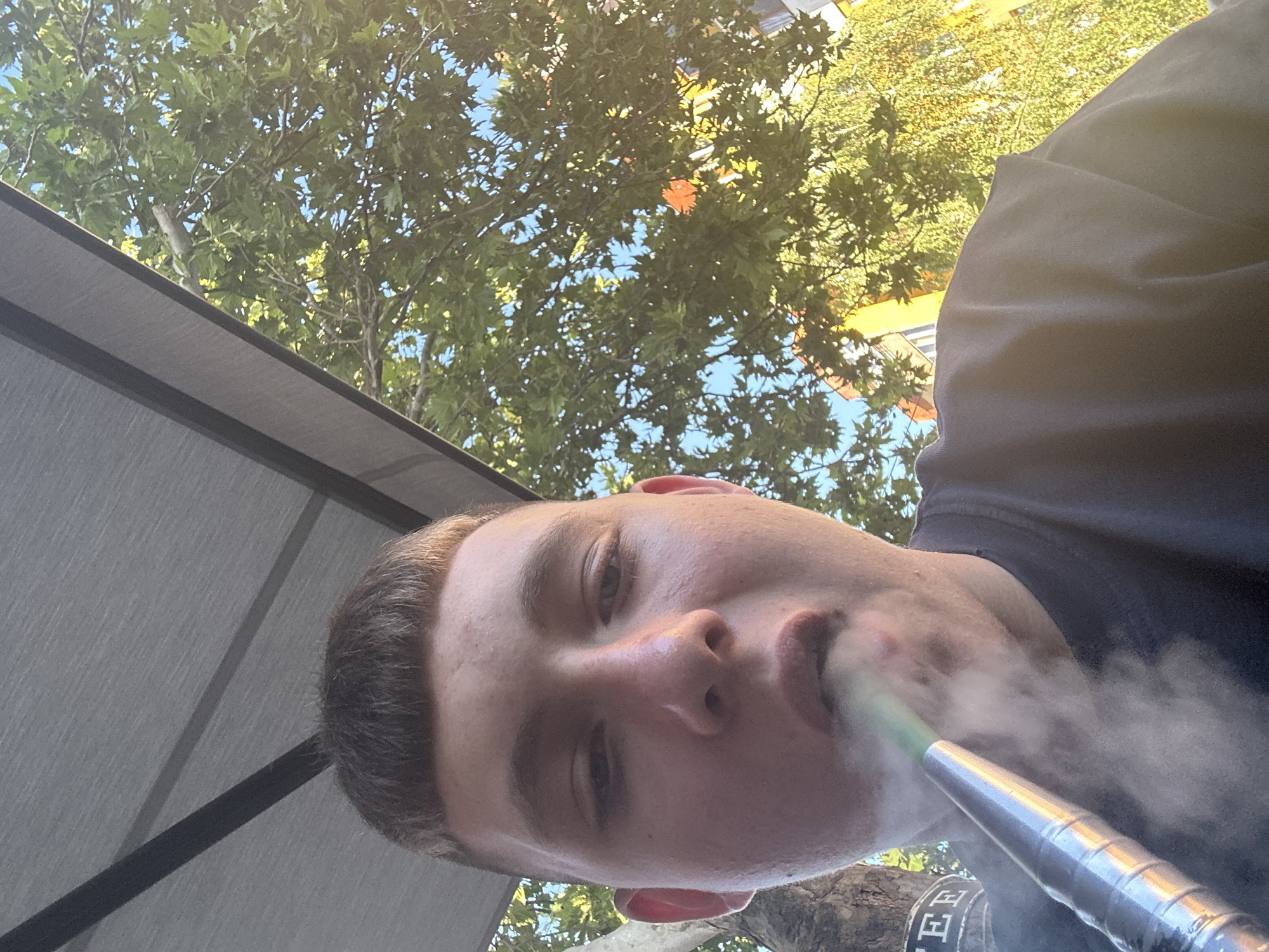 A young person with a short haircut is taking a selfie outdoors while smoking a hookah. They are wearing a dark t-shirt and the image is taken from a low angle looking up. In the background, lush green trees with sunlight filtering through them are visible, along with parts of buildings. The person's face is close to the camera, and smoke from the hookah is obscuring their mouth and chin. A small portion of a sign with the letters "FEE" is visible to the left. The setting appears to be a casual outdoor area in a city, possibly a cafe or a street with trees.