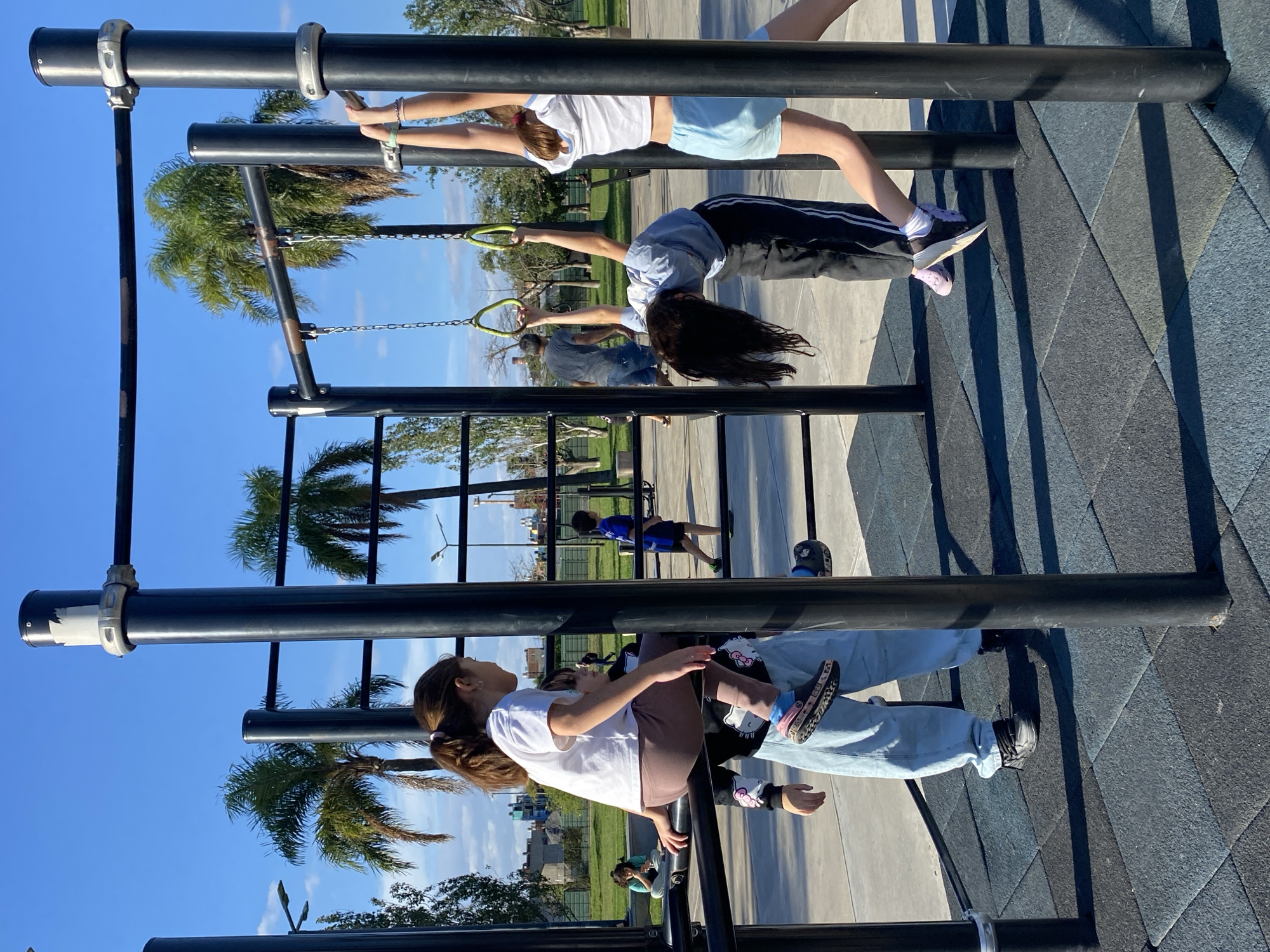 The image shows three young people using outdoor exercise equipment in a park. The equipment consists of parallel bars and gymnastic rings. Two girls are hanging on the bars and rings, performing exercises. One girl is sitting on a bar. The park has a cushioned surface and palm trees in the background. In the distance, other people are visible, including a child running. The location appears to be outdoors in a sunny area. Based on the vegetation and architectural styles that are visible, this may be in Buenos Aires, Argentina.