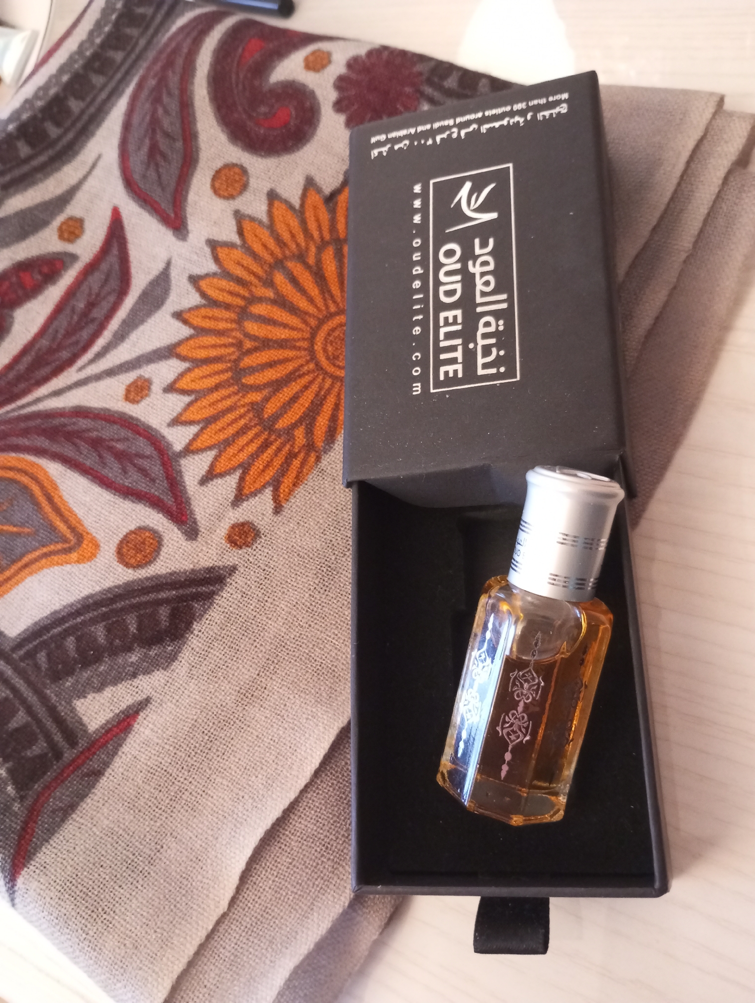 The image shows a bottle of Oud Elite perfume in its black box. The perfume bottle is small and has a silver cap. The label on the box indicates the brand and the company's website. The box is open, revealing the dark interior. To the left of the box, there is a piece of patterned fabric with orange, brown, and gray colors. Another folded piece of light gray fabric is seen to the right of the box. The background is a light surface, likely a table. There's no indication of the city of Chaikovsky, Russia in the image.