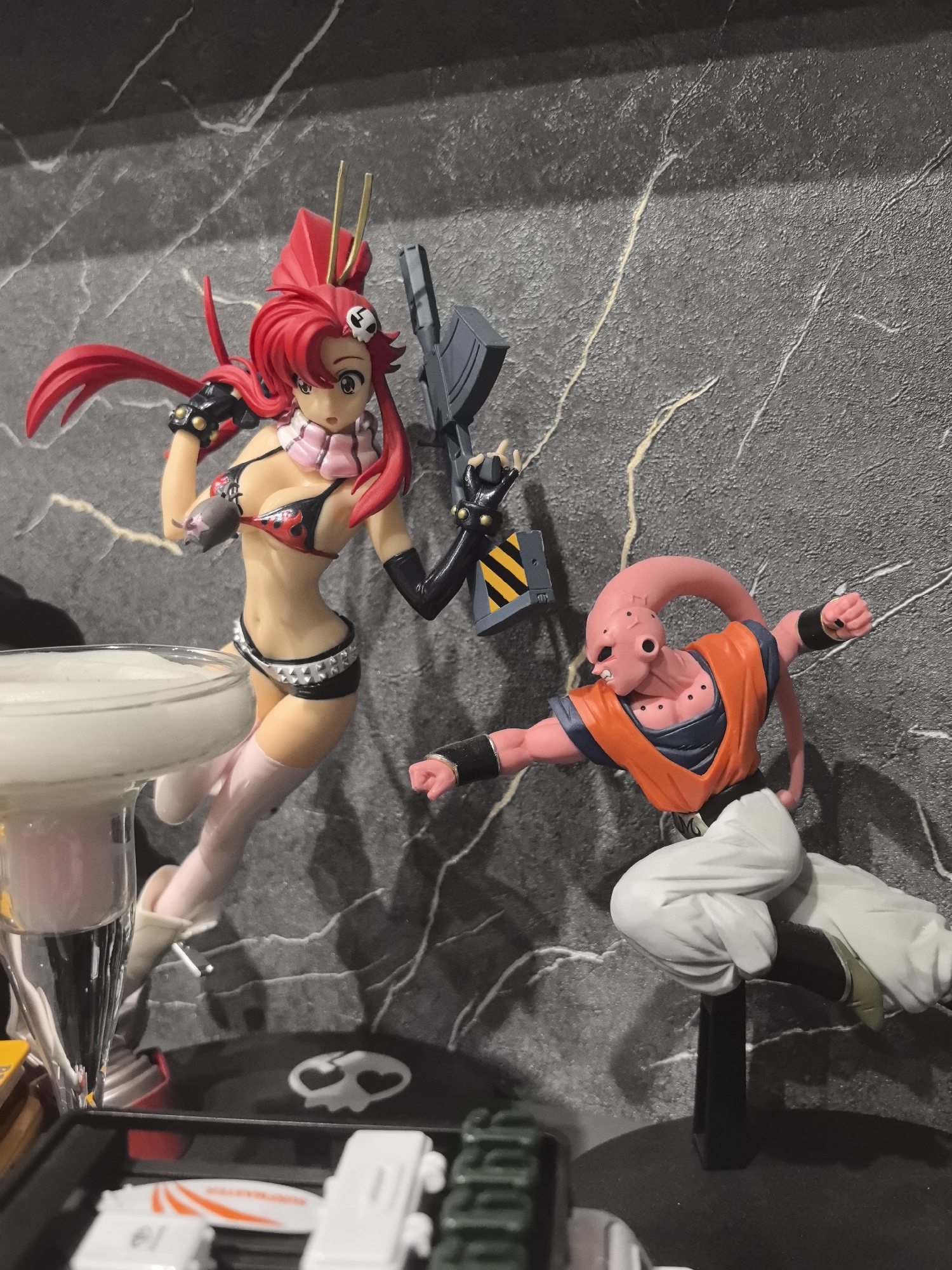 The image shows two anime action figures against a dark, textured background that resembles a marble wall with white and yellow veins. On the left is a female character with long, bright red hair styled into a large, spiky bun and flowing strands. She wears a black bikini with red flames, a pink scarf, studded black briefs, and thigh-high pink stockings. She has black gloves and holds a gray rifle. On the right is a pink, alien-like character with large ears and dark spots on its face and chest. It wears an orange vest over a dark blue shirt and loose white pants. Both figures appear to be in dynamic poses, suggesting a battle or action scene. In the foreground, partially visible, are parts of other objects, including a clear glass object on a stand, and several small, toy-like vehicles or components. The lighting is even, highlighting the figures and their details. There is no identifiable information about the location of Amursk, Russia in the image.