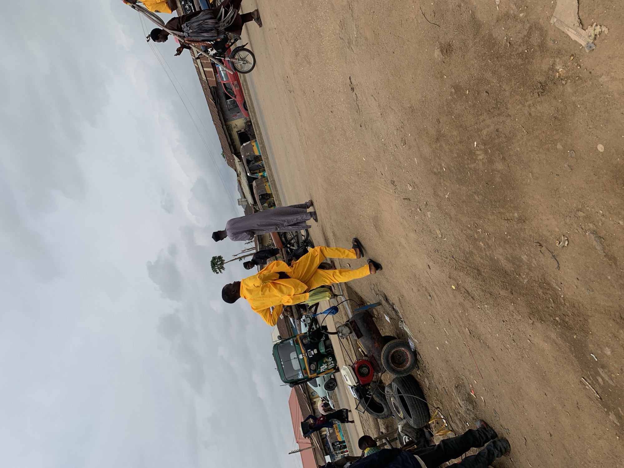The image captures an outdoor street scene in Rafin Par, Nigeria, under an overcast, cloudy sky. The primary feature is an unpaved, dusty, light brown dirt road that dominates the lower two-thirds of the frame.
Several individuals are visible, engaged in various street activities. In the mid-ground, a man in a striking bright yellow full-body outfit stands beside the road, looking towards the right. Around him are assorted vehicle parts, including loose tires and what appears to be a small engine or generator, suggesting a roadside repair or maintenance spot. A green motorized tricycle (often called a Keke Napep) is parked nearby. Further down the road, another man, dressed in a long grey traditional garment and a cap, walks away from the camera. A person pushing a bicycle can be seen further in the background. Partial views of other people are scattered throughout the scene.
Along the dusty road, several vehicles are present, including multiple tricycles and a partially visible red car. Buildings with corrugated iron roofs and simple facades line the street in the distance, indicating commercial or residential structures.
The time of day appears to be daytime, with a uniformly grey and cloudy sky covering the upper portion of the image. The scene portrays a typical bustling street environment, characterized by local transport, pedestrians, and informal roadside commerce in a rural or semi-urban Nigerian setting. No legible text is discernible on any signs or objects within the frame.