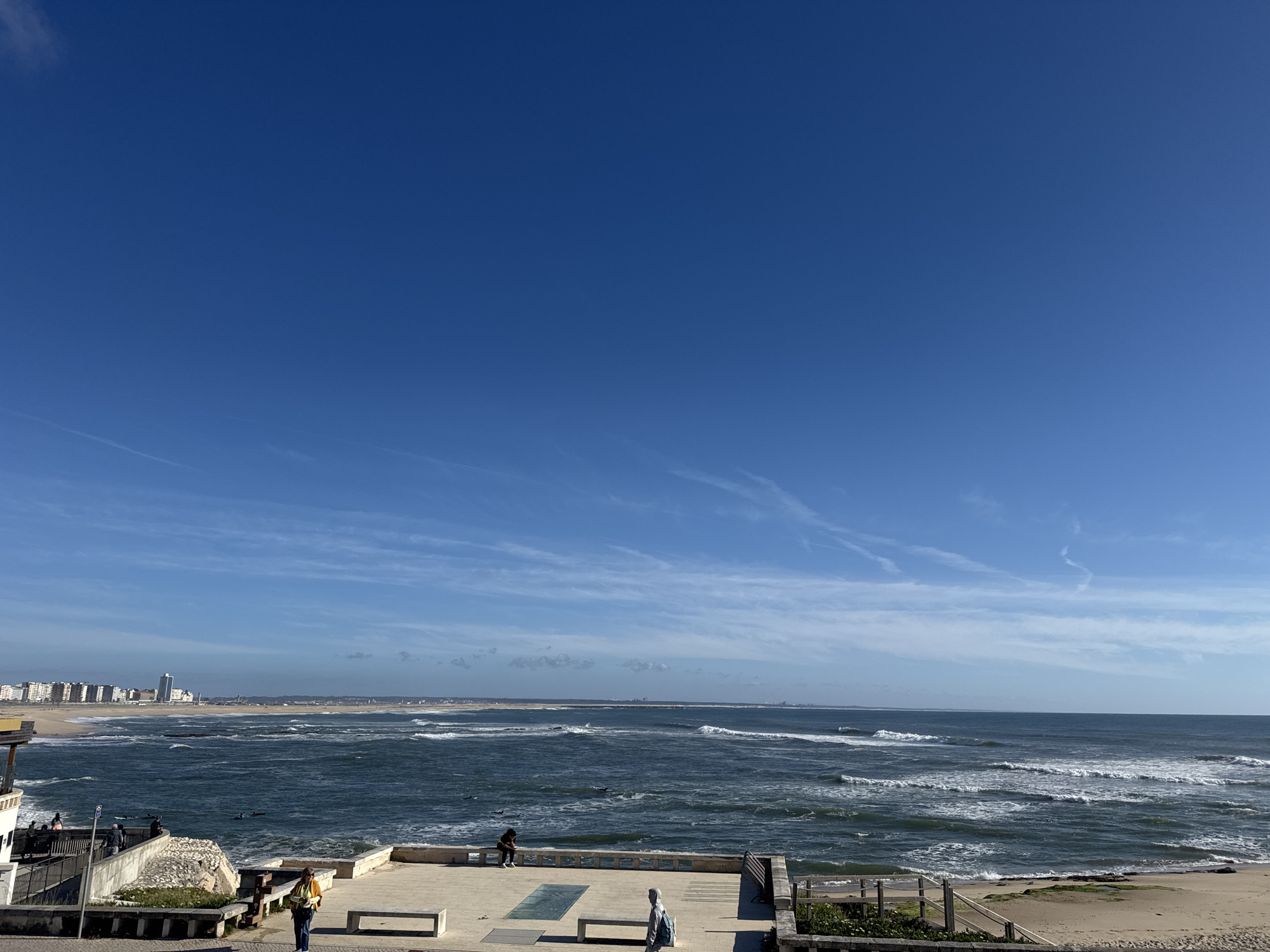 The image depicts a coastal scene in Figueira da Foz, Portugal. The foreground features an elevated concrete platform with benches and a low railing. Several people are present: a woman walking, a person sitting on a ledge, and a person standing. Below the platform is a rocky area with some vegetation. The middle ground shows the Atlantic Ocean with moderate waves breaking along the coastline. Beyond the beach is a landmass, possibly a peninsula, with some structures visible. In the background, the city skyline of Figueira da Foz is discernible, including high-rise buildings. The sky is clear blue with thin, wispy clouds in the middle portion.