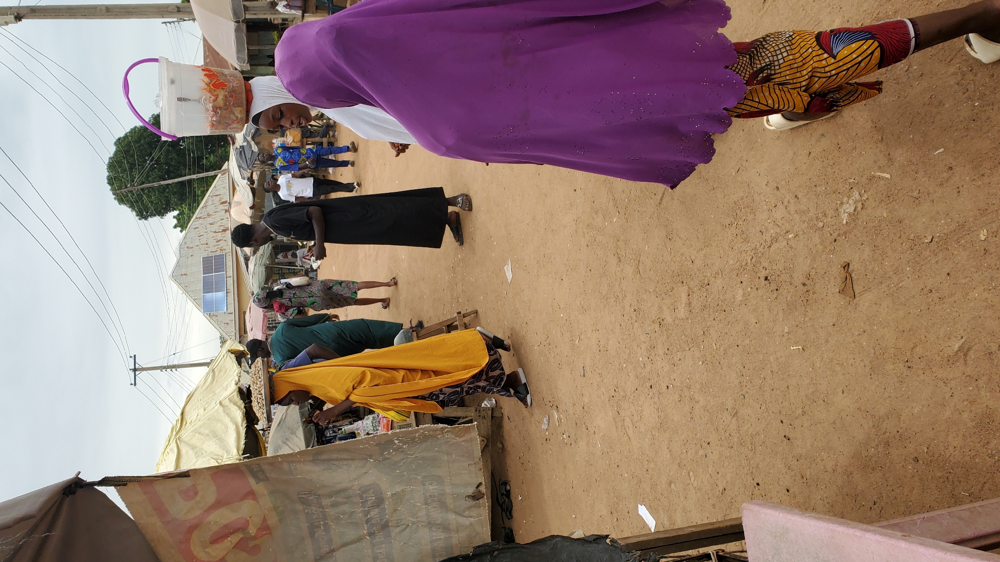 The image depicts a bustling outdoor market scene in Jalingo, Nigeria. A woman wearing a bright yellow headscarf and robe walks from left to right across a sandy path, carrying a woven tray of food balanced on her head. She is walking past a stall with a tan awning. Behind her, a man in a dark robe and another in a green robe are also present. Further back, several other individuals are visible, some walking and others standing, suggesting a busy market environment. A white bucket containing food and a spoon is balanced on the head of a person in the foreground, partially obscured by a purple veil and patterned leggings. The background shows buildings with corrugated metal roofs, some adorned with solar panels, indicating a blend of traditional and modern infrastructure. Overhead, utility poles and wires crisscross the sky. The ground is unpaved and sandy, typical of many market areas in Nigeria.