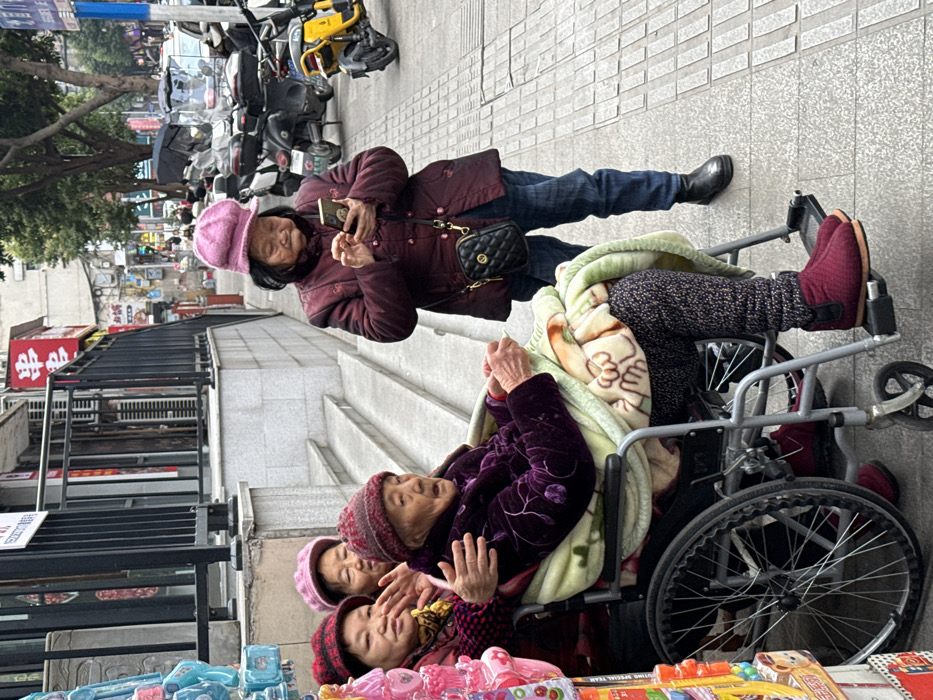 Four elderly Chinese women are present on an urban sidewalk in Jiangjin, China. In the foreground-left, an elderly woman with a dark red knit cap and purple velvet jacket is seated in a silver manual wheelchair, covered by a light green blanket with cartoon motifs. Her hands rest on the blanket. An elderly woman in a red knit cap and dark red jacket stands to her immediate left, embracing the woman in the wheelchair with her right arm around the seated woman's left arm. A third elderly woman, wearing a light pink knit cap and dark jacket, stands partially obscured behind the embracing woman, looking forward. To the right, an elderly woman in a pink newsboy cap, maroon jacket, blue jeans, and black ankle boots stands facing slightly left, operating a gold-colored smartphone with both hands. She wears a small black quilted shoulder bag. The sidewalk features a textured paving pattern. In the background, a building with a black metal railing and concrete stairs rises. Chinese characters are visible on red signage ("串串"). Further back, a bustling street features multiple parked motorcycles and scooters, including a yellow model, and trees.