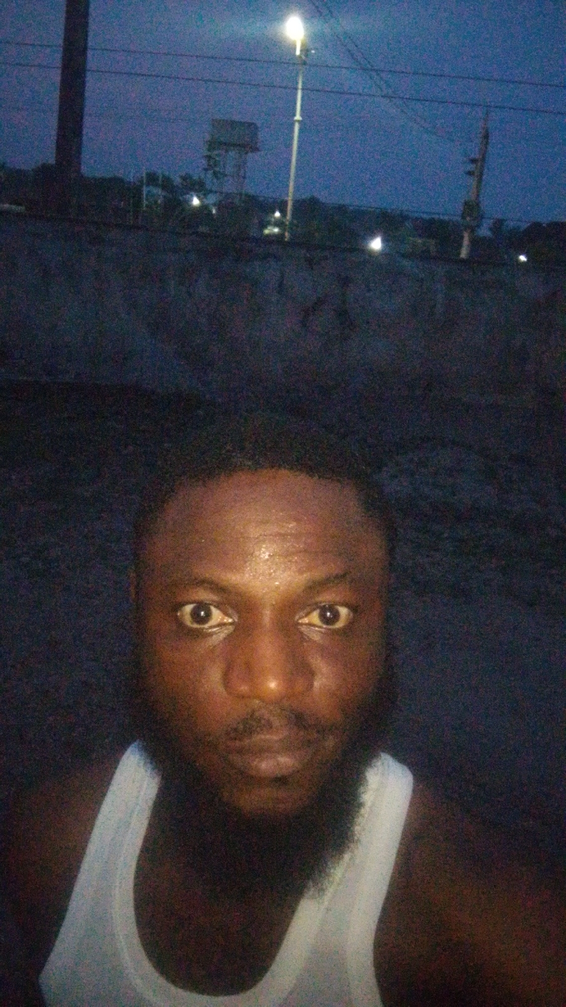 A selfie of a Black man with a beard, wearing a white tank top. The man has dark skin and a sweaty forehead. In the background is a street at night, illuminated by a streetlight. A water tower is visible in the distance. The location appears to be Lokoja, Nigeria.