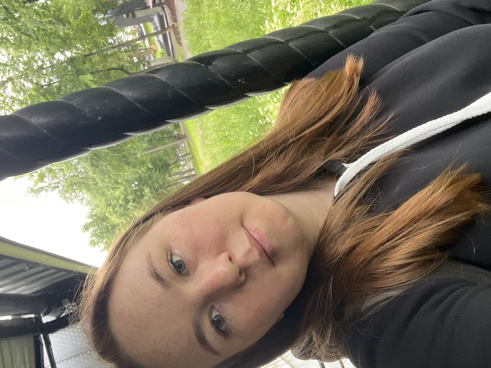 A close-up selfie shows a young woman outdoors in Chaikovsky, Russia. She has long, reddish-brown hair, styled loosely around her face and shoulders, and light-colored eyes that engage directly with the camera. Her complexion exhibits subtle freckles, and she wears a calm, neutral expression, with a hint of a slight upward curve to her lips. She is dressed in a black hoodie, part of which is visible, featuring a white drawstring detail on the front.
A thick, black, twisted rope-like structure, possibly a railing, beam, or part of a playground apparatus, dominates the upper and central portions of the image, running diagonally. This object appears sturdy and industrial in design.
The background behind the woman and the black structure features vibrant green foliage, indicative of trees and grass, suggesting a park, garden, or natural public area. The lighting is bright but diffused, suggesting daytime conditions, possibly overcast or with indirect sunlight. The image captures a personal moment in a tranquil outdoor setting. No text is visible in the media file.