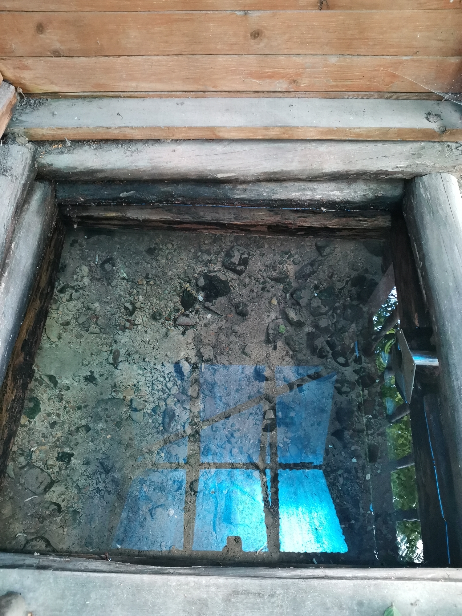 This is a top-down, close-up, eye-level shot of a shallow, man-made well or water feature, likely outdoors. The well is constructed of rough-hewn, weathered wood posts and beams, forming a square or rectangular opening. The water within is clear and shallow, revealing a bed of small, grey stones and sediment. A bright blue tarp or plastic sheet is submerged in the water, creating a reflective surface that distorts the view of the stones and the surrounding environment. The area above the well is framed by wooden planks, suggesting it is covered or part of a larger wooden structure. The overall impression is one of a naturalistic or rustic setting, perhaps in a park or garden.