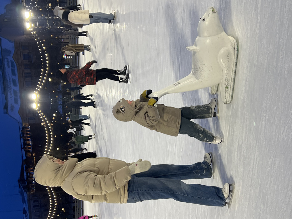 An outdoor ice rink scene is depicted at dusk or early night, located in Konnyushennaya Square, Saint Petersburg, Russia. The environment is illuminated by numerous overhead string lights, creating a festive canopy, and by tall streetlights. Classical-style multi-story buildings with snow-dusted rooftops are visible in the background, characteristic of a historic city square.
In the foreground, an adult individual, dressed in a beige hooded puffer jacket, blue jeans, and white mittens, stands on white ice skates, facing slightly right. Adjacent to them, a young child wearing a beige hooded coat with an animal face design (possibly a squirrel or cat) on the hood, blue-green pants, yellow gloves, and white ice skates is actively pushing a white, seal-shaped ice skating aid. The child's mouth is open. The aid features a vertical handle and shows signs of wear.
In the midground and background, multiple other individuals are scattered across the ice rink, engaged in recreational ice skating. Most are clad in winter jackets, hats, and gloves. One notable figure in the midground is a male wearing a black hat and a red camouflage-patterned jacket, skating from right to left. The ice surface is visibly marked by skate activity.