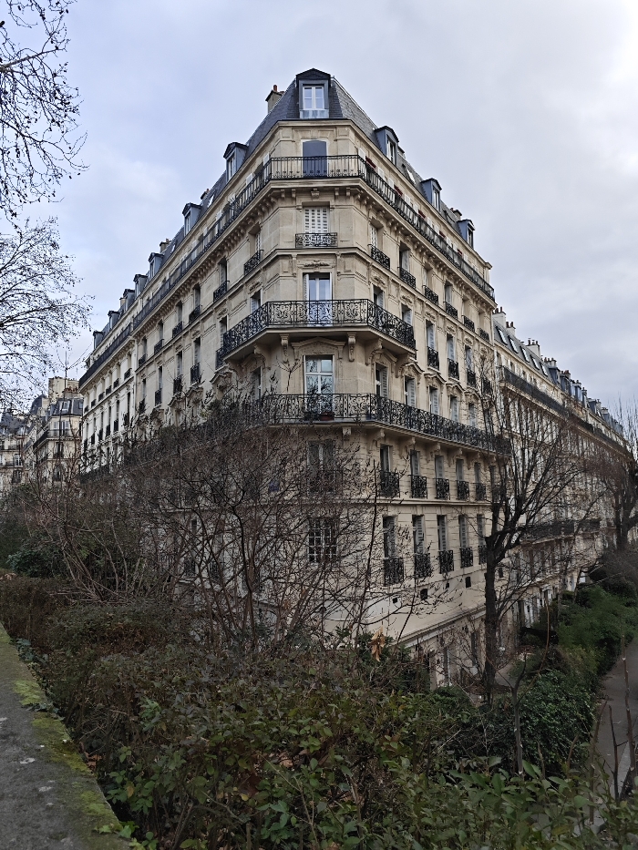 The image displays a prominent, multi-story Haussmannian-style building located at a street corner in Paris, France, captured under an overcast sky. The structure is built with light-colored stone and features six primary floors, surmounted by a dark slate mansard roof with multiple dormer windows. Its facades are lined with numerous rectangular windows, many of which are fitted with closed white louvered shutters. Black wrought-iron balconies are present on several levels, with a particularly ornate, continuous balcony wrapping around the building's second story. Similar Parisian architectural styles are visible extending along both intersecting streets in the background. The foreground is occupied by a dense urban green space, consisting of various evergreen bushes and numerous bare deciduous trees and shrubs, indicating a cool season. A moss-covered stone retaining wall defines the left edge of this green space, and a paved surface, likely a road or pathway, is partially visible to the far right. No individuals, activities, or interactions are discernible within the frame.