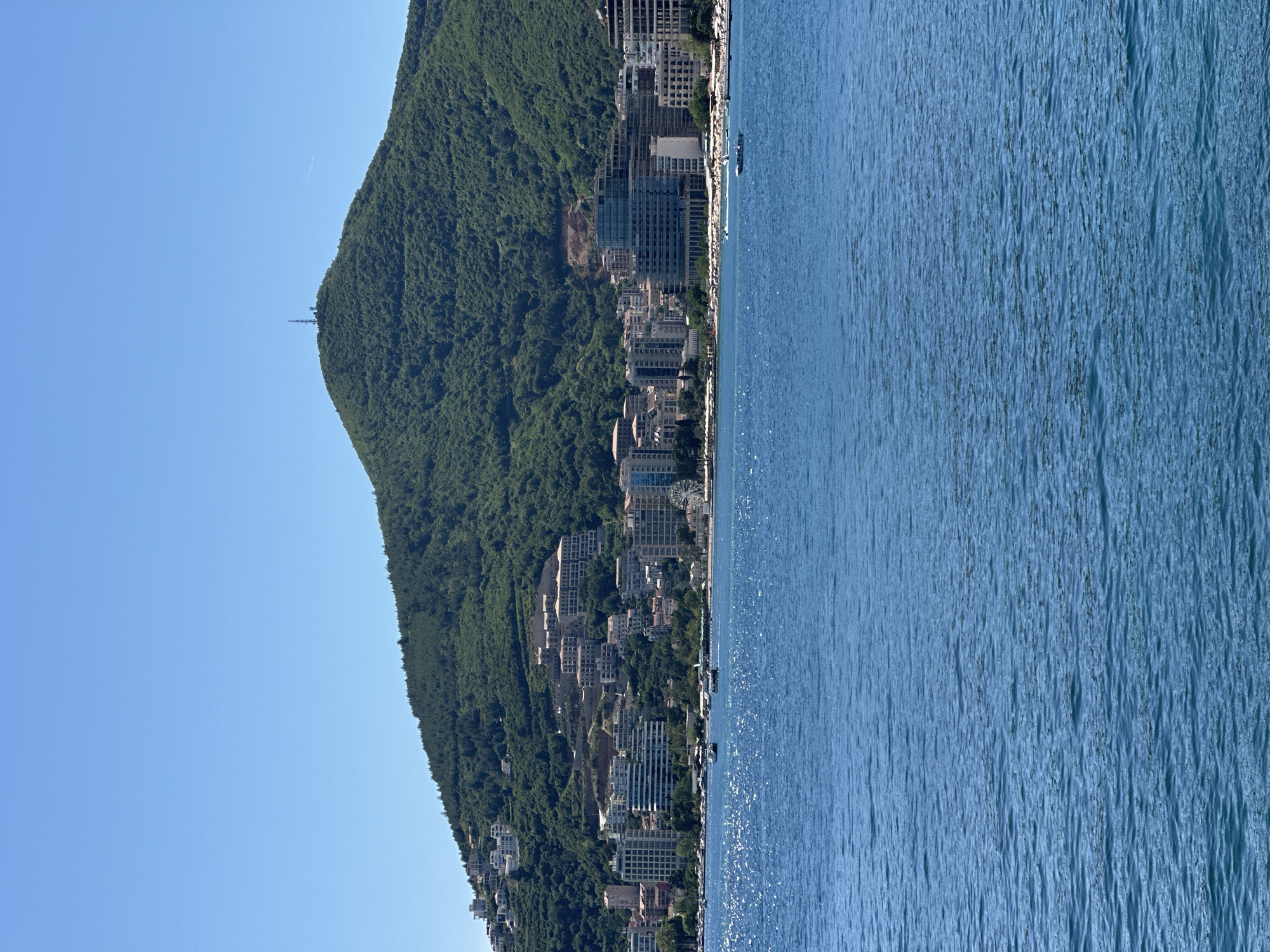 The image depicts a coastal scene in Budva, Montenegro. On the left, a steep, verdant mountain dominates the landscape, with residential buildings clustered along its lower slopes. These buildings vary in size and architectural style, suggesting a mix of housing options. A communication tower stands atop the mountain's peak. To the right of the mountain, a body of water stretches out, its surface shimmering with sunlight. The sky is clear and light blue. Overall, the scene showcases the juxtaposition of natural and urban elements.