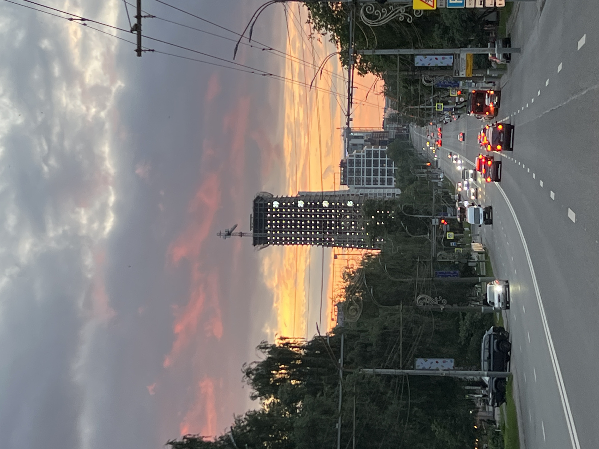 The image depicts a sunset over a city street in Izhevsk, Russia. The sky is filled with dramatic clouds, tinged with pink and orange hues from the setting sun. A tall, unfinished skyscraper with numerous lights is prominently visible in the mid-ground, dominating the skyline. The street itself is busy with traffic, with numerous cars and a bus navigating the roadway. Trees line the sides of the street, and overhead power lines crisscross the sky. The overall atmosphere is urban and dynamic, capturing the transition from day to night in a bustling city.