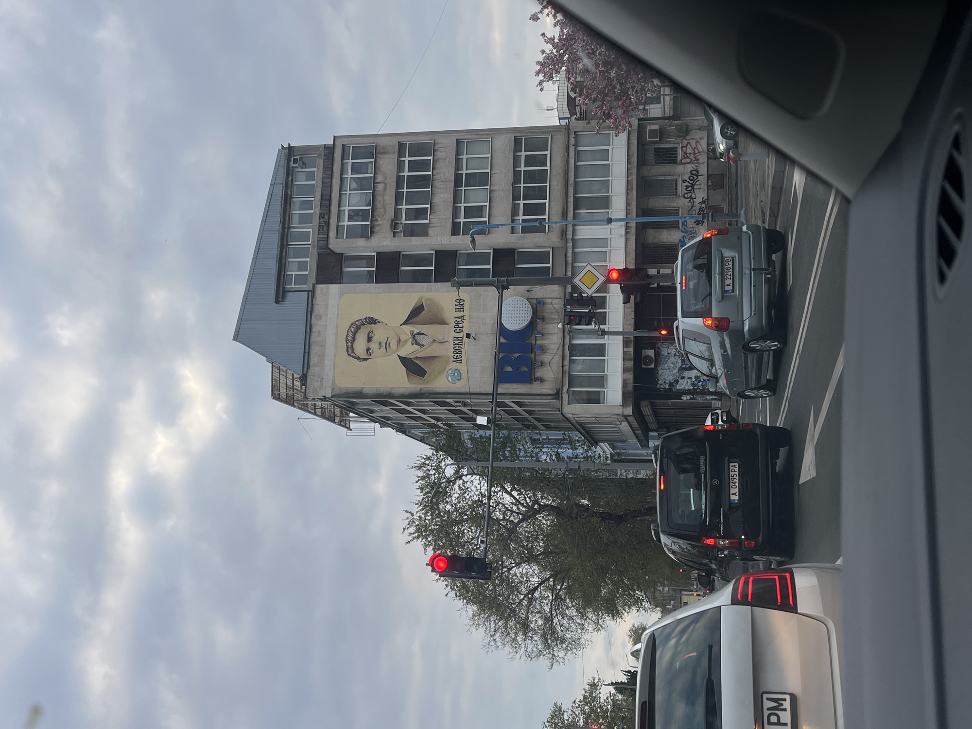 The image captures a street scene in Burgas, Bulgaria, from the perspective of someone inside a car. The view is dominated by a multi-story building on the left, featuring a large mural of a young man, likely Vasil Levski, a national hero. Next to the mural, Cyrillic text reads "Левски сред нас" (Levski among us/within us), and below it, a blue sign displays "BIG".
In the foreground, the interior of the vehicle, including part of the dashboard and a vent, frames the scene. The weather appears overcast with a cloudy sky. On the road, several cars are stopped at a red traffic light. Two red lights are clearly visible, one in the mid-ground and another further down the street. A yellow diamond-shaped sign, indicating a priority road, is also visible near the traffic lights.
Specific cars in the traffic line include a silver SUV with license plate A 2298 PB, and a black van with plate A 0495 PA, both indicating registration in the Burgas region. The rear of a white car with plate A 1481 PM is also visible in the immediate foreground. A prominent tree with green foliage stands between the road and the buildings, and some pink blossoms are visible on a tree or bush in the upper right corner, suggesting spring. The building itself is made of concrete, with numerous windows, and shows some wear and tear, including visible air conditioning units and graffiti on adjacent structures. The overall impression is that of a bustling urban environment on a cloudy day.