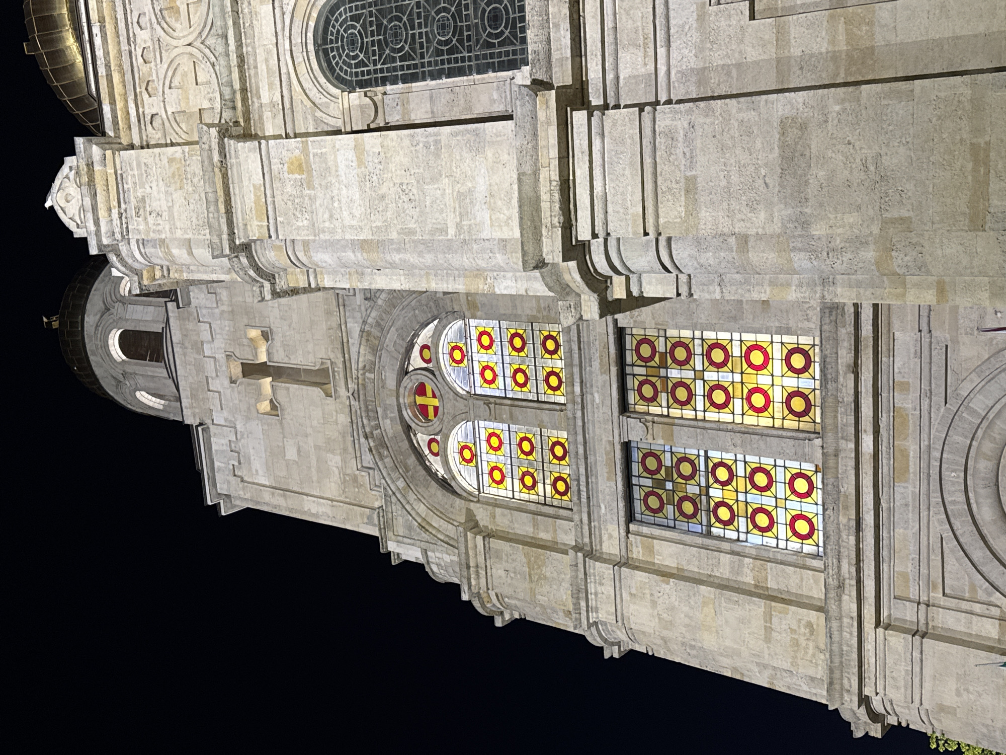 The image presents an illuminated section of a grand, ornate stone building at night, likely a cathedral or church, set against a dark sky. The architecture appears classical or neoclassical, characterized by light-colored stone blocks, intricate carvings, columns, and cornices.
A prominent feature is the array of windows. On the right side of the main vertical structure (when the image is viewed upright), there are multiple rectangular stained-glass windows, each composed of smaller square panes. Each square pane contains a distinct geometric pattern: a central red circle surrounded by a yellow ring, set against a lighter yellow background. To the left of these rectangular windows is a large, circular stained-glass window. Its central pane features a cross in red and yellow, flanked by similar red and yellow circular patterns found in the other windows.
Further to the left and slightly below this circular window, a carved stone cross is integrated into the building's facade. Above the stained-glass windows, a large round window with intricate dark tracery is visible. The building's surfaces are heavily detailed with decorative elements, including rounded moldings and pilasters, casting deep shadows that emphasize their three-dimensional form under the artificial lighting.
The time of day is clearly night, with the building brightly illuminated, making the vibrant colors of the stained glass stand out. No people or activities are visible, and there is no discernible text on the building. The image provides a detailed view of the architectural grandeur, typical of significant historical structures in locations like Varna, Bulgaria.