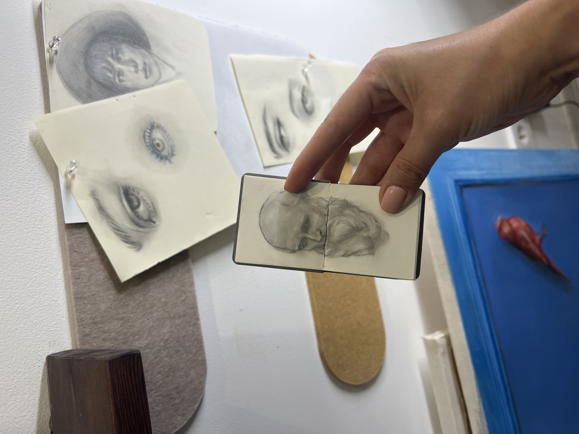 A hand is holding a small sketchbook with a drawing of a bearded man on its cover. The sketchbook is propped up against a wooden easel, and behind it are several other drawings of faces and eyes, pinned to a wall with clear pushpins. To the left of the drawings, a dark wooden block is mounted on the wall. Below the drawings, a bright blue framed canvas with a red bird painting is visible. The background is a textured white wall.