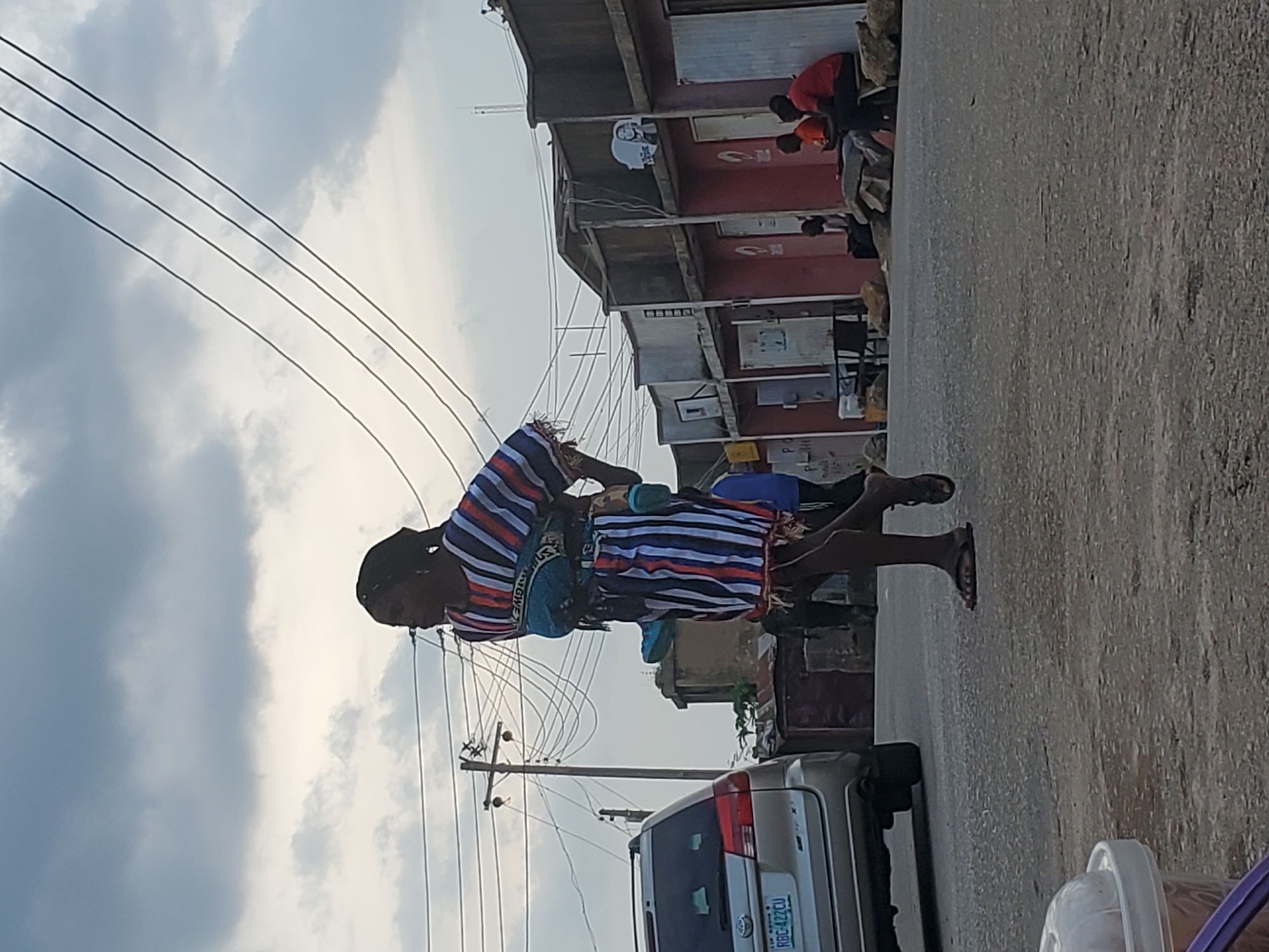 The image depicts a street scene in Jos, Nigeria, with a person walking along a paved road. The person is wearing a striped dress and carrying a bag. In the background, there are buildings with red facades and corrugated iron roofs. A car is parked on the side of the road. The sky is cloudy, suggesting it might be a cloudy day. Power lines are visible overhead. The overall impression is of a typical day in a busy urban environment. The scene appears to be captured during daylight hours. No specific emotions are discernible from the image. There is text visible on the license plate of the car, which reads "RBC 422CU".
