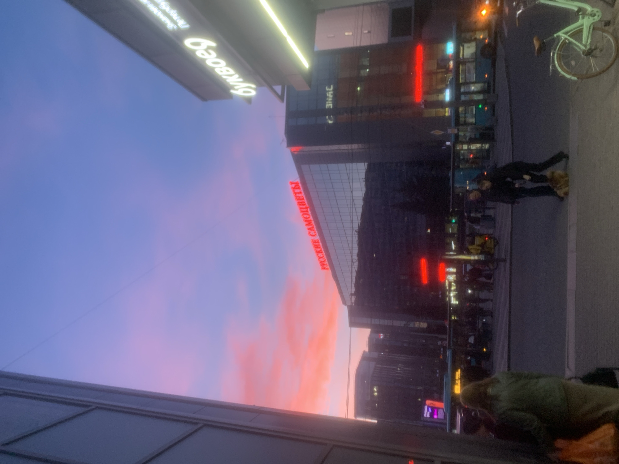 The image captures an urban street scene in Saint Petersburg, Russia, during twilight or early evening. The sky dominates the upper left, transitioning from a deep purple-blue at the top to a vibrant pink and orange sunset glow towards the horizon, indicating a clear sky.
Several buildings line the street, illuminated by artificial light. On the left, a building features a prominent, bright neon sign displaying "Буквоед" (Bukvoed), a well-known Russian bookstore chain. Another sign, partially obscured above it, appears to be "РИВ ГОШ" (Rive Gauche), a cosmetics retailer. A long, bright yellow-green light strip runs horizontally above these signs.
A large, modern building occupies the center-right, its glass facade reflecting the colorful sky. A prominent red neon sign on this building reads "РУССКИЕ САМОЦВЕТЫ" (Russkie Samotsvety), indicating a jewelry store. Below it, the letters "ЗНАС" are faintly visible, likely part of another sign. Further to the right, a vertical red neon light illuminates another section of the building or a nearby structure.
The lower right portion of the image shows a paved street or sidewalk. Two individuals are standing in the mid-ground on the sidewalk; one person wears dark clothing, and the other has a lighter, possibly fluffy, accessory. In the immediate foreground, another person wearing a green jacket and light-colored pants is visible, appearing to be bent over or looking down. A light green bicycle is parked on the sidewalk in the upper right. In the background, a bus or trolleybus with illuminated windows can be seen. The scene depicts a typical city environment as day transitions into night, with various neon signs providing light and commercial cues.