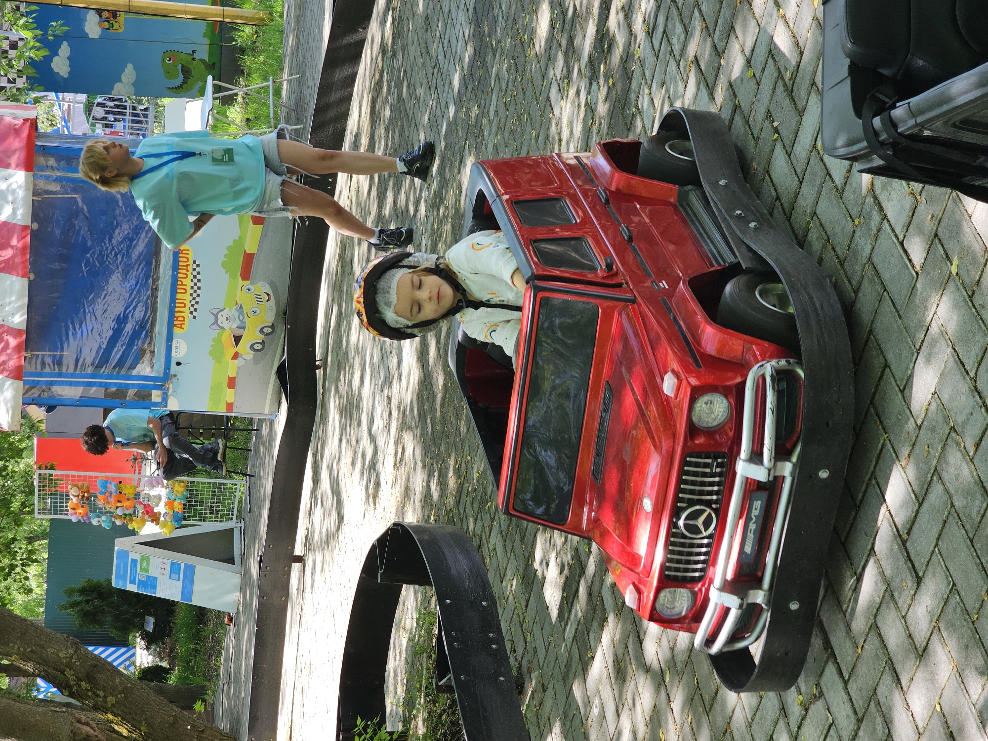 Captured in Khimki, Russia, the scene depicts a child resting in a red, toy-sized AMG Mercedes G-Wagon, designed for coin-operated rides, within an outdoor park or recreational area, confirmed by the backdrop of trees and shaded, brick-paved ground. The child, wearing a helmet, appears to be sleeping or resting with eyes closed. A woman with light hair is seen near a carnival-style booth featuring cartoon graphics; another person sits near a display of stuffed animals.