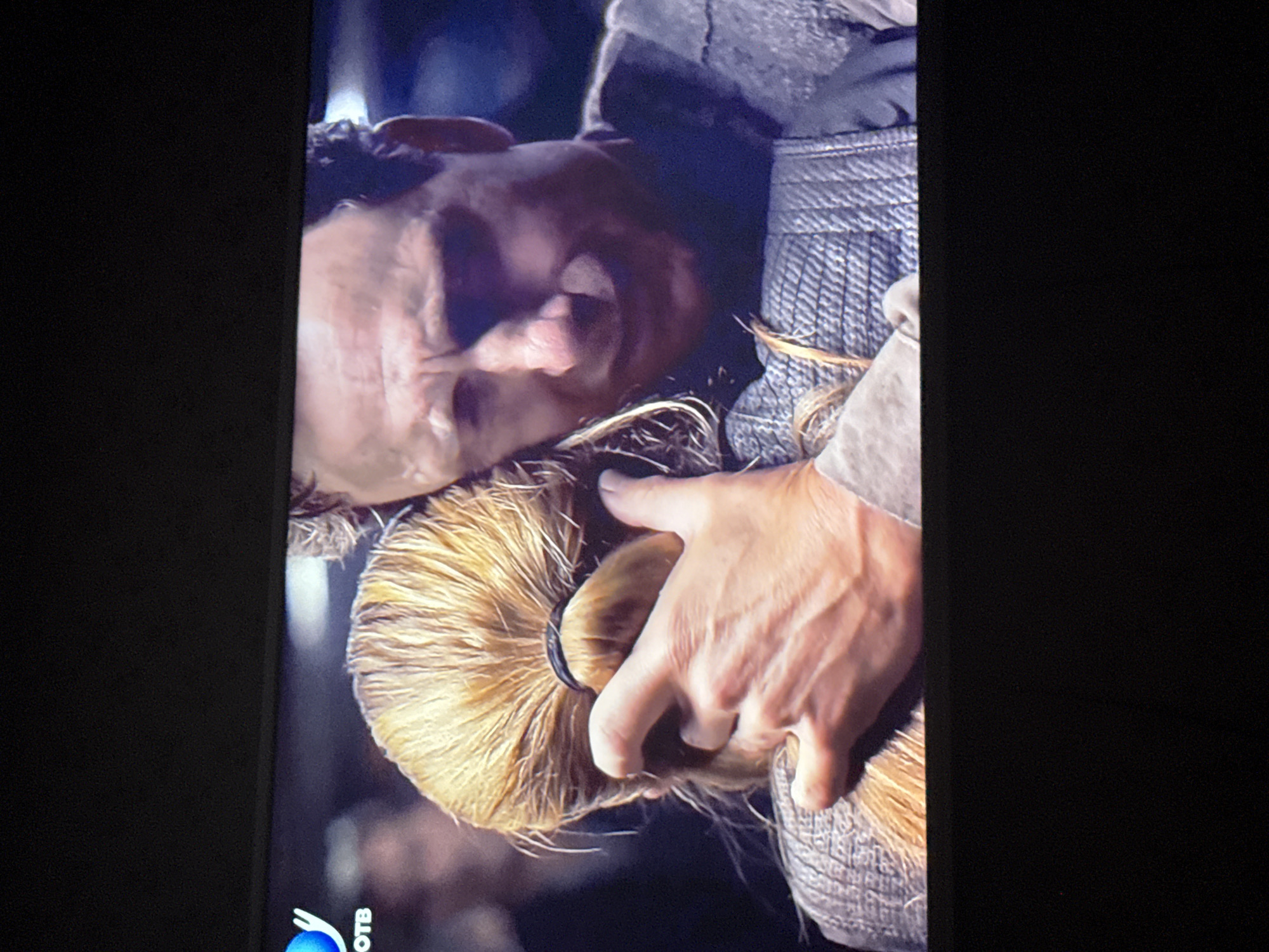 A man with wrinkled skin is lying down while another person is touching his blonde ponytail. The person touching the ponytail is wearing a gray woven garment. The location is not discernible from the image.