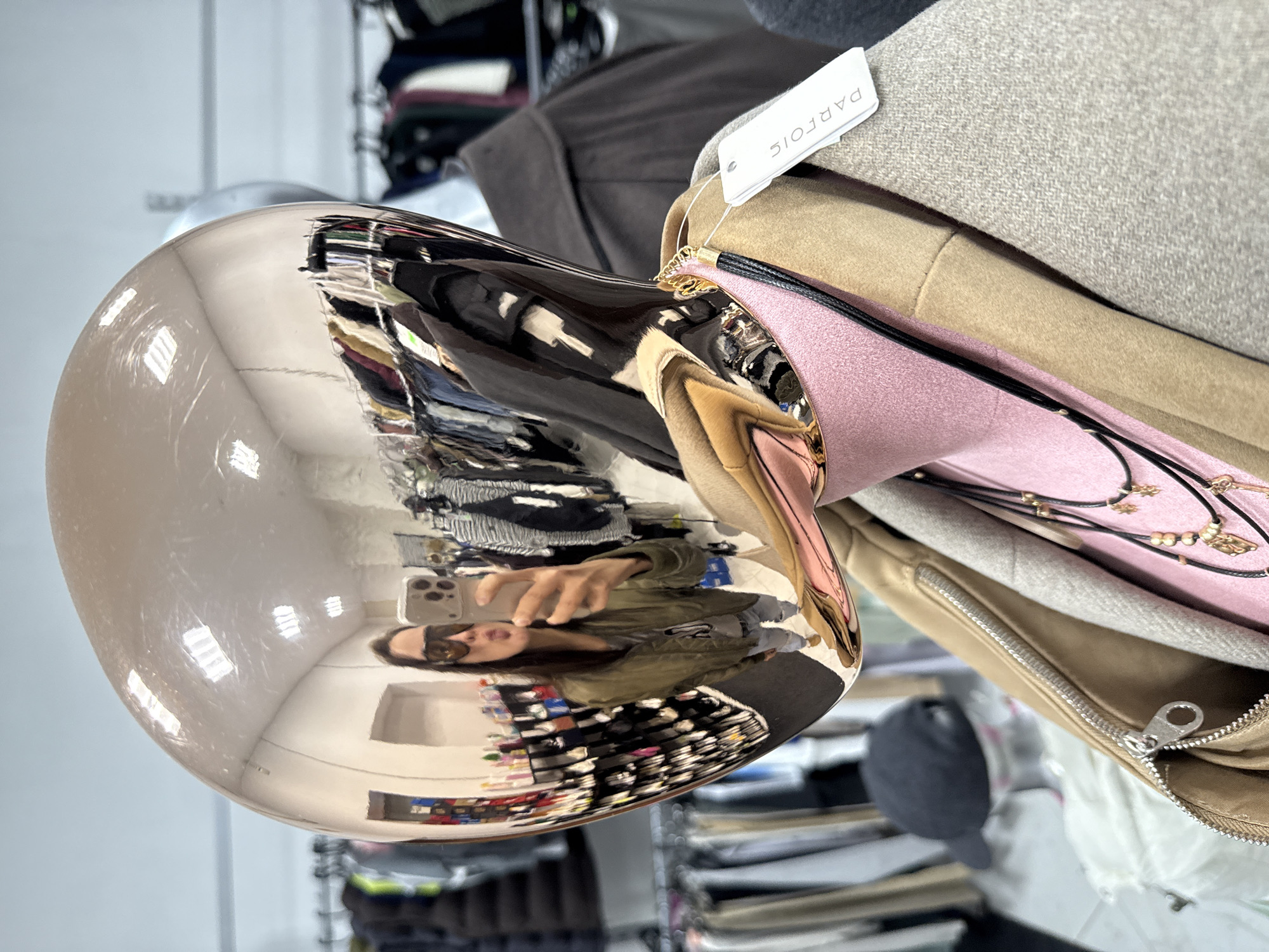 The image depicts a distorted reflection in a shiny, metallic, spherical object, likely a decorative item within a clothing store. The reflection shows a woman wearing sunglasses holding a smartphone, presumably taking the photo. The store's interior is visible in the reflection, revealing racks of clothes and merchandise. To the right of the sphere are stacked layers of clothing, including garments in pink, tan, and grey. A white tag with the word "PARFOIS" is attached to one of the garments. The general environment suggests a retail setting. There is no information to indicate that this photo was taken in the city of Chaikovsky, Russia.
