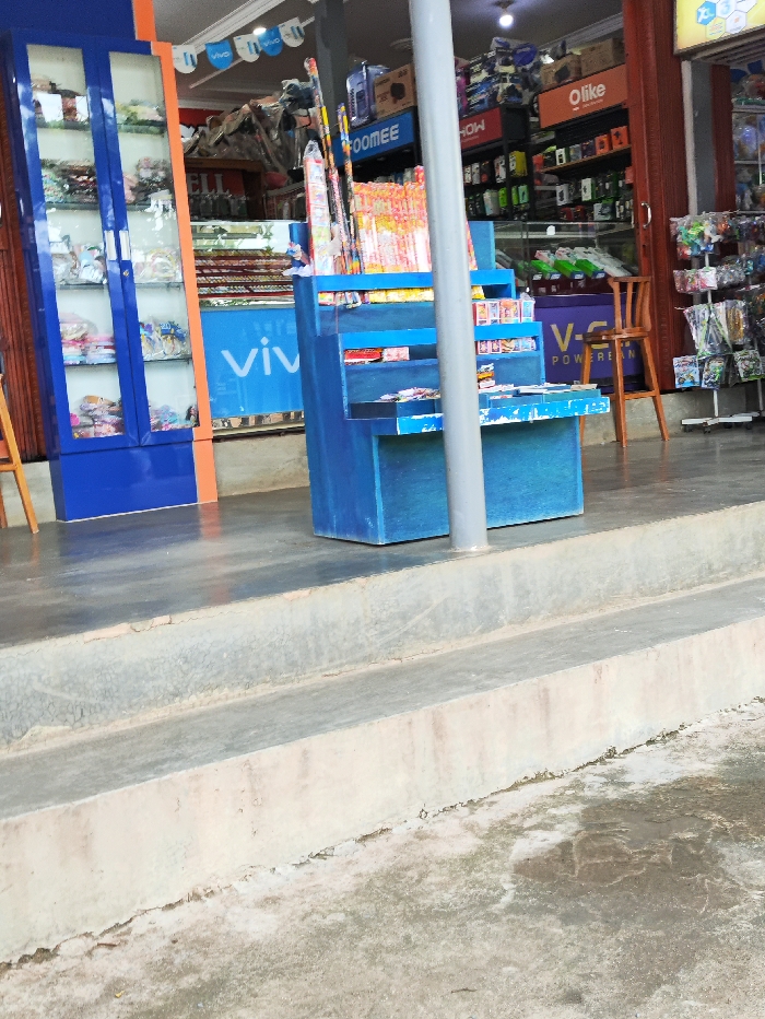 This image depicts the entrance area of a retail shop, likely in Kecamatan Selagai Lingga, Indonesia. The scene is devoid of people.
In the foreground, a concrete ground transitions into two low concrete steps leading up to the shop's entrance. The ground and steps show some wear and staining.
The shop entrance features several prominent objects and displays. To the left, a tall, blue, glass-fronted display cabinet holds various packaged goods, possibly hair accessories or small trinkets, neatly arranged on shelves. Next to it, a large light blue sign with the white "vivo" logo is visible.
In the center of the frame, a multi-tiered blue display stand holds numerous items. The top tiers are filled with what appear to be fireworks or sparklers, while lower shelves contain small packaged products, possibly matches or other small items. A white pole runs vertically through the middle of this display.
Further inside the shop, visible in the background, are shelves stocked with various electronics accessories. Brands like "FOOMEE," "HOW," and "Olike" are identifiable on signs above these shelves. Another blue sign reads "V-G POWER BANK." To the right, a single brown wooden chair is placed, and a white rack displays packaged toys or other colorful merchandise. Banners with the "vivo" logo are strung across the shop's ceiling area.
The lighting suggests it is daytime, with interior lights also illuminated. No specific activity is occurring, but the setup indicates a functioning retail business.
