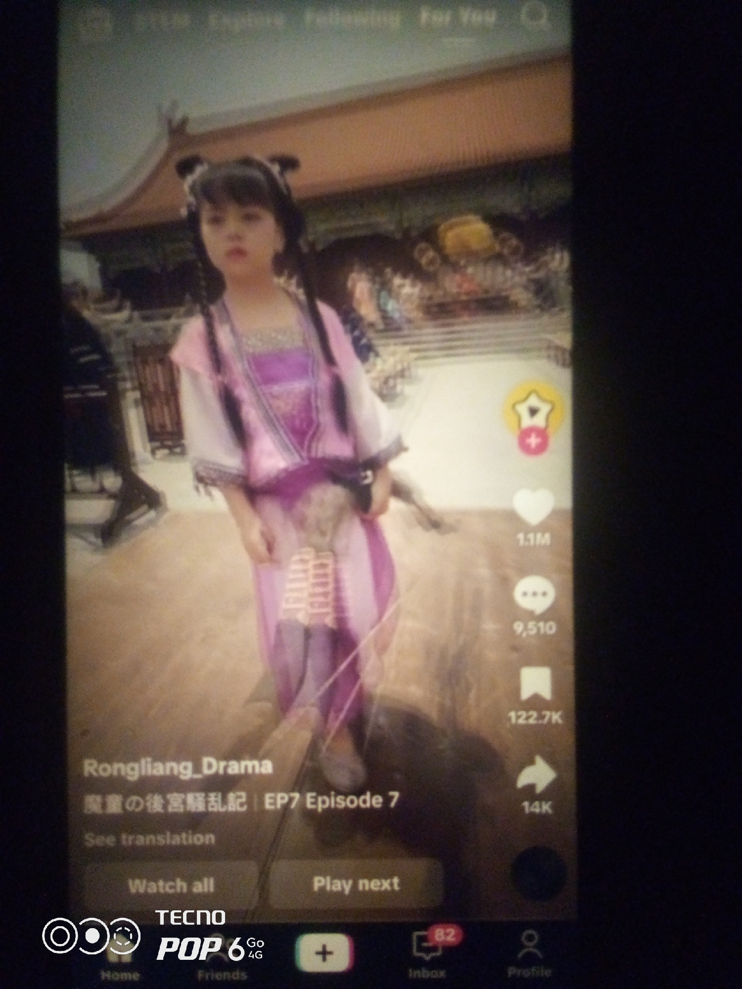The image shows a screenshot of a TikTok video. A young person in traditional East Asian attire, possibly a historical costume, is the central subject. They have their hair styled in two buns and are wearing a purple and pink layered dress with ornate details. The background appears to be a set designed to resemble traditional East Asian architecture, with a reddish-brown roof. On the right side of the screen, there are common TikTok interface elements: a star icon with a plus sign, a heart icon labeled "1.1M", a speech bubble icon labeled "9,510", a bookmark icon labeled "122.7K", and a share icon labeled "14K". At the bottom of the screen, text overlays indicate the video title as "Rongliang_Drama" followed by Japanese characters and "EP7 Episode 7". Below this, a "See translation" button is visible, along with "Watch all" and "Play next" buttons. The bottom navigation bar of the TikTok app shows icons for "Home", "Friends", "Inbox" (with a notification count of "82"), and "Profile". The lower-left corner of the screenshot also displays the "TECNO POP 6 Go 4G" branding, indicating the device on which the screenshot was taken. The image is blurry, suggesting it was taken with a phone camera in low light or with motion. There is no indication of the city of Howayi, Nigeria in the image.