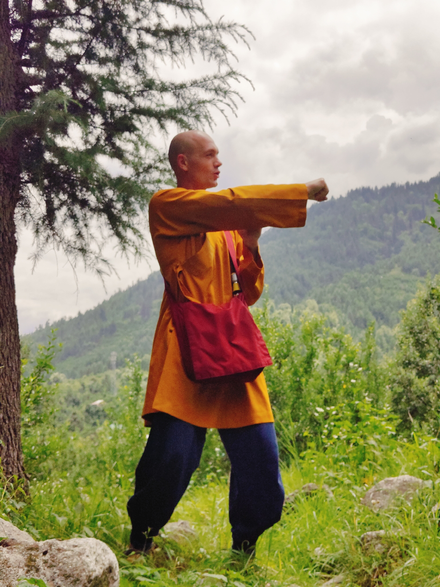 A bald man, wearing a yellow robe and blue pants, stands in a forest, practicing martial arts. He is holding his right arm outstretched, with his fist clenched. A red bag is slung across his left shoulder. The man is facing the right, with his head turned to the left. The background is a lush green forest with a mountain range in the distance. The sky is cloudy and overcast. The location is the city of Bahang, India.