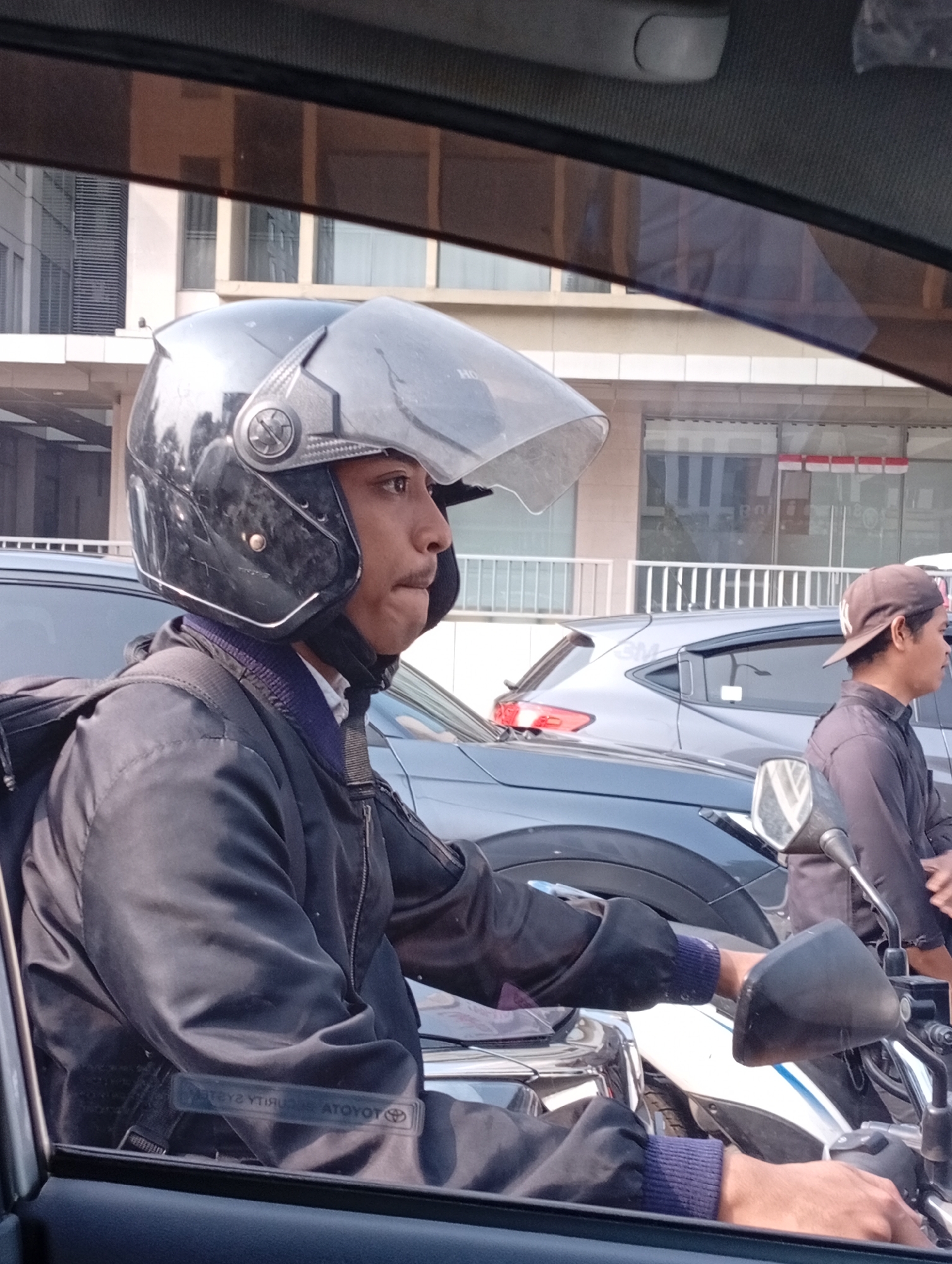 The image shows a man wearing a black helmet and jacket, riding a motorcycle in what appears to be a city street in Lengkong Kulon, Indonesia. The man is looking to his right. In the background, other vehicles, including cars and another motorcycle, are visible, along with buildings. The scene is captured from the perspective of someone inside a car, looking out. The lighting suggests it is daytime.