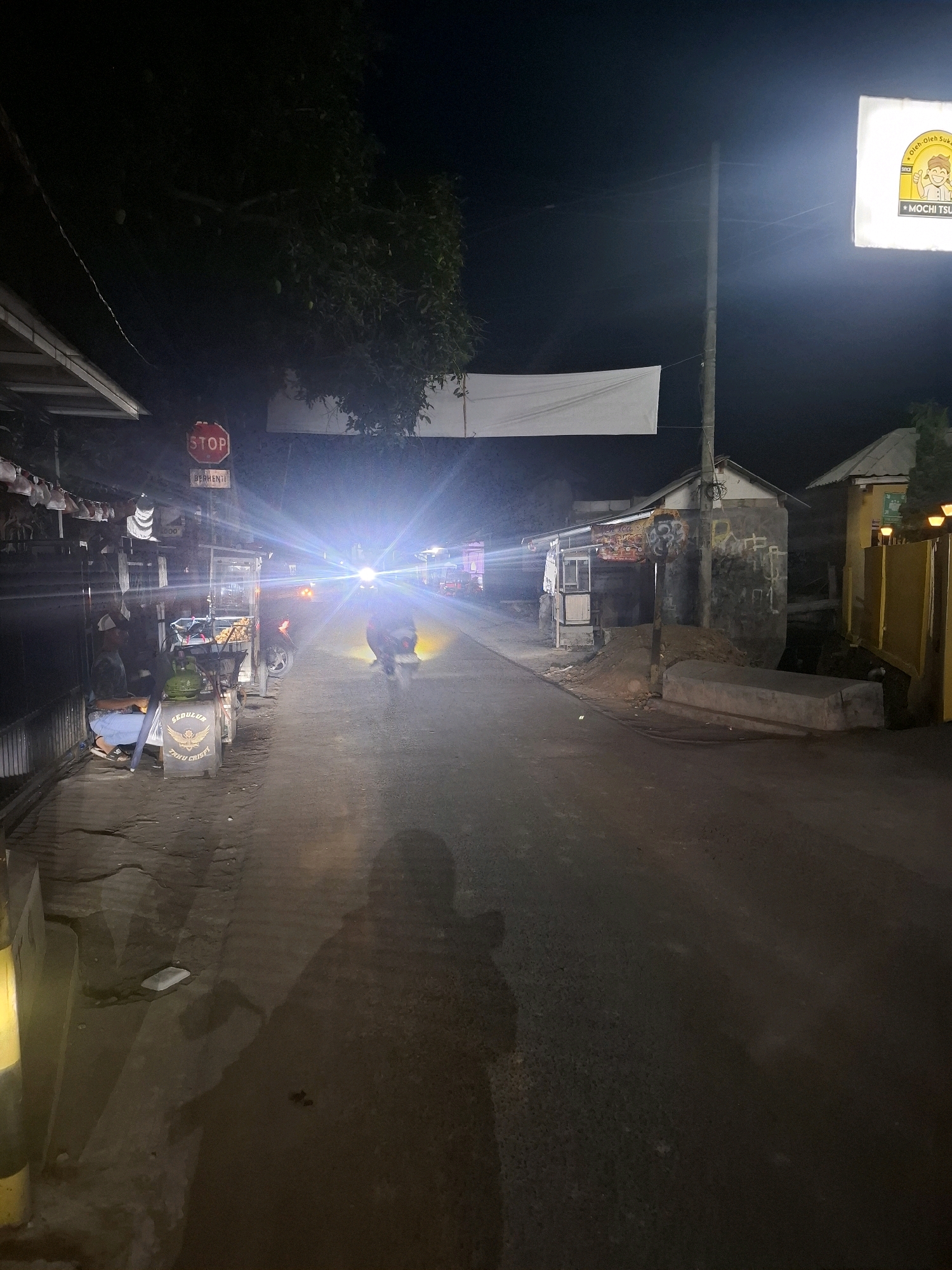 In Karangtengah, Indonesia, at night, a street scene unfolds under artificial lights. A motorcycle with its headlights on, illuminating the road, is moving away from the viewer. A person is seated on the left side of the road, near a food cart. The food cart appears to be selling fried food, as indicated by the signage that reads "GEDULUR TAHU CRISPY". A red STOP sign is visible on a pole on the left side of the road, with the word "BERHENTI" below it. A banner or cloth is strung across the street in the background, and a lit sign with a cartoon character and the text "MOCHI TSU" is visible on a pole to the right. The environment is a moderately lit street with buildings and trees lining the sides. The asphalt road is visible, and shadows are cast on it.