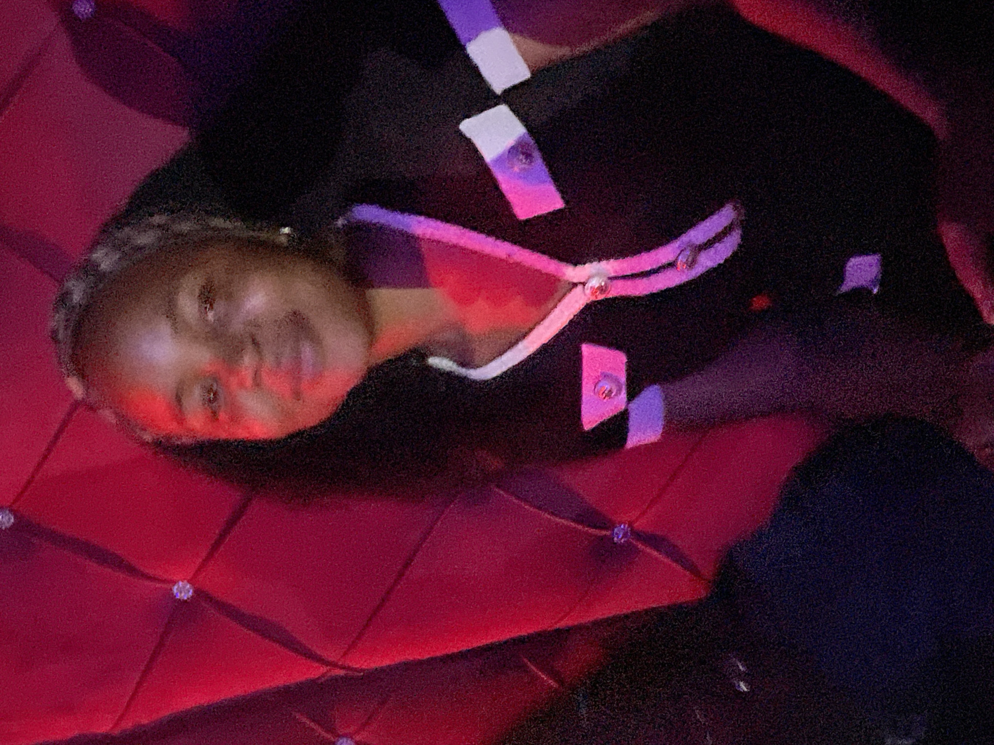 The image shows a young Black woman with braided hair sitting in a dimly lit room with red upholstered seating. She is wearing a black dress with white and purple trim. The background consists of tufted red velvet upholstery with visible diamond-shaped patterns and some embedded rhinestones. The lighting is primarily red, casting shadows and highlighting certain areas of her face and clothing. The overall impression is of an indoor, possibly dimly lit, social setting. No specific location in Durumi, Nigeria can be identified from the visual information provided.