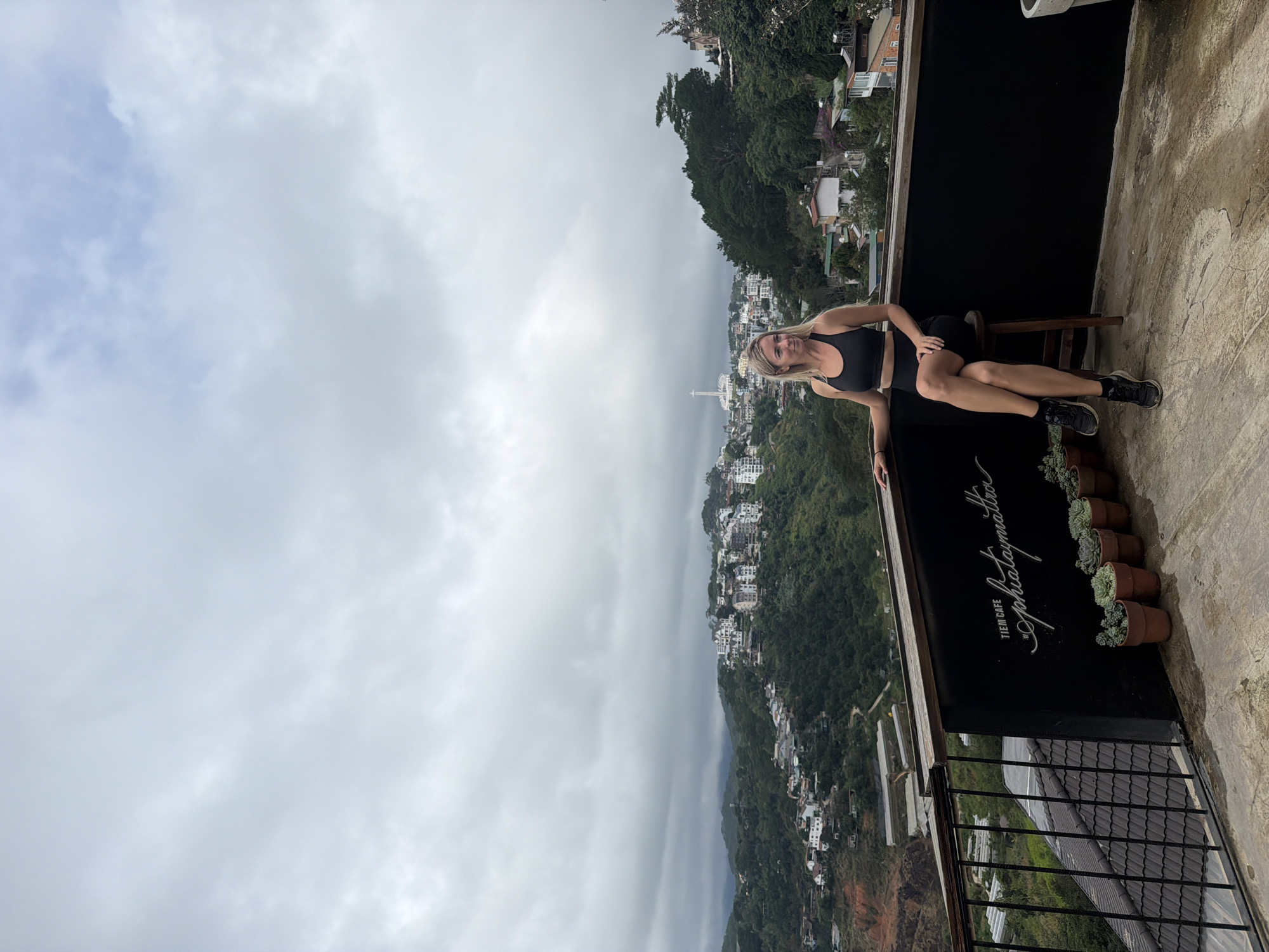 A blonde woman wearing a black sports bra and black shorts is sitting on a wooden stool on a balcony overlooking a hillside city. The city has many white buildings with red roofs, nestled amongst lush green trees. The sky is overcast with gray clouds. The balcony has a black railing and a black wall with white text that reads "TIEM CAFE sophiatmattra". There are several potted plants on the balcony floor. The woman is looking to her right and has her legs crossed.