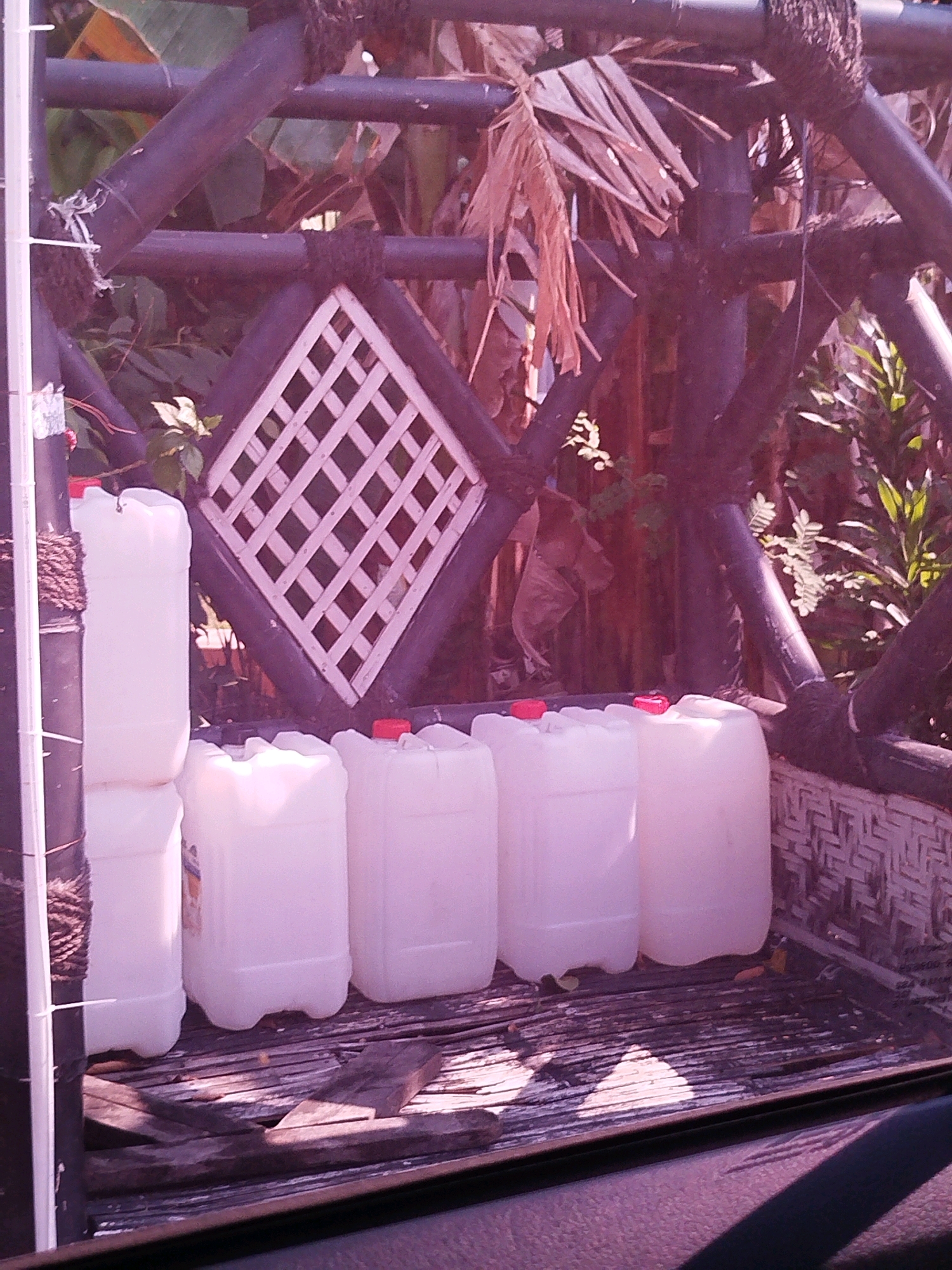The image shows several white plastic jerrycans with red caps, neatly arranged on a wooden surface. The jerrycans appear to be new and are positioned in what looks like the bed of a pickup truck or a similar platform. Behind the jerrycans is a rustic wooden fence structure with diagonal beams and woven elements. In the background, there are dry leaves and green foliage, suggesting an outdoor environment. The lighting indicates it is daytime, with sunlight casting shadows. The overall scene has a slightly aged or weathered appearance, particularly the wooden structures. The image does not contain any people or any identifiable landmarks specific to Cagakagung, Indonesia.