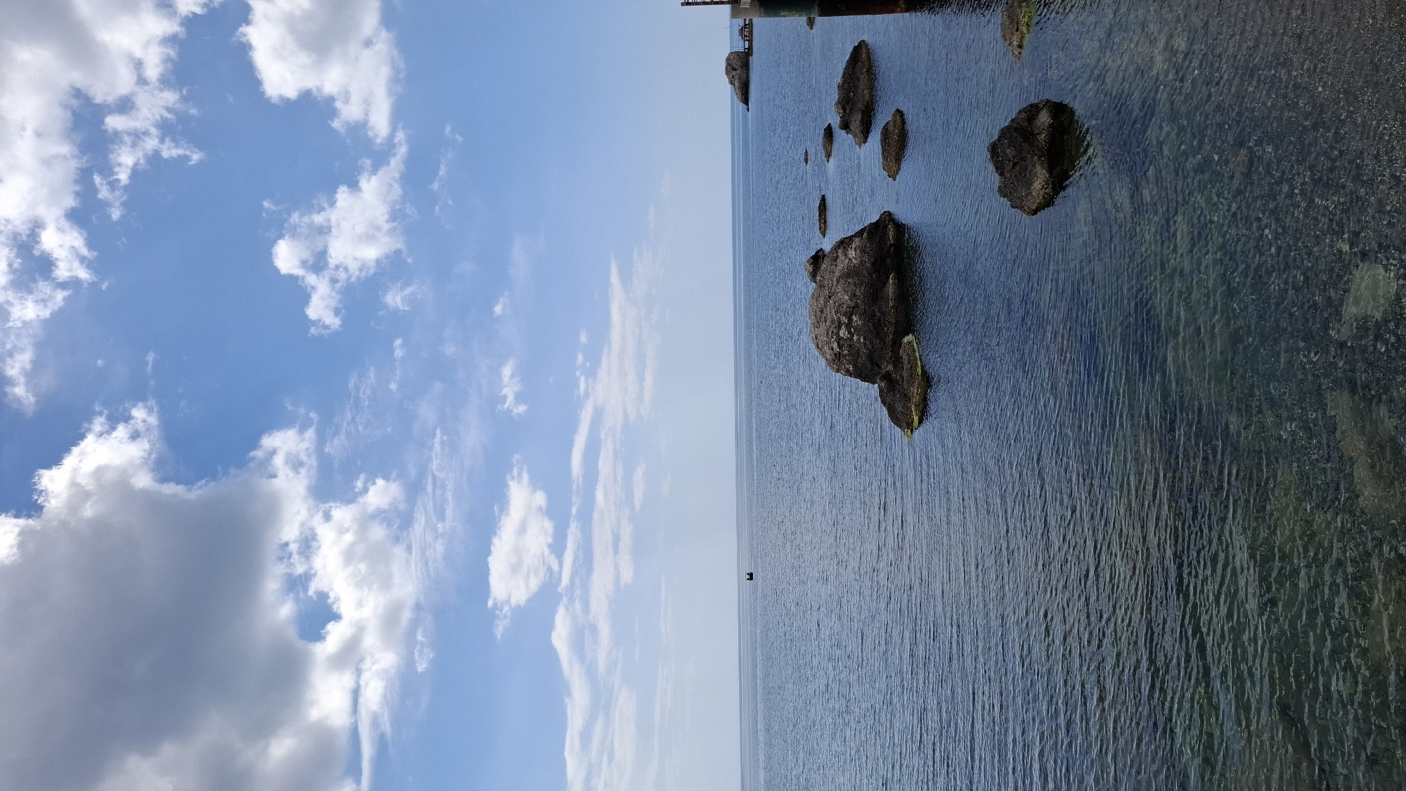 The image captures a seascape scene. The sky, predominantly blue with scattered white clouds, occupies the left side of the image. On the right, the sea displays varying shades of blue, reflecting the sky above. Several rocks of different sizes are visible in the water. Their dark color contrasts with the sea around them. The water appears clear, with the seabed visible in some areas. In the distance, a dark, indistinct structure is present, likely a pier or a building near the coast. A small black object floats in the middle of the sea. This image was taken in the city of Unknown, Unknown.
