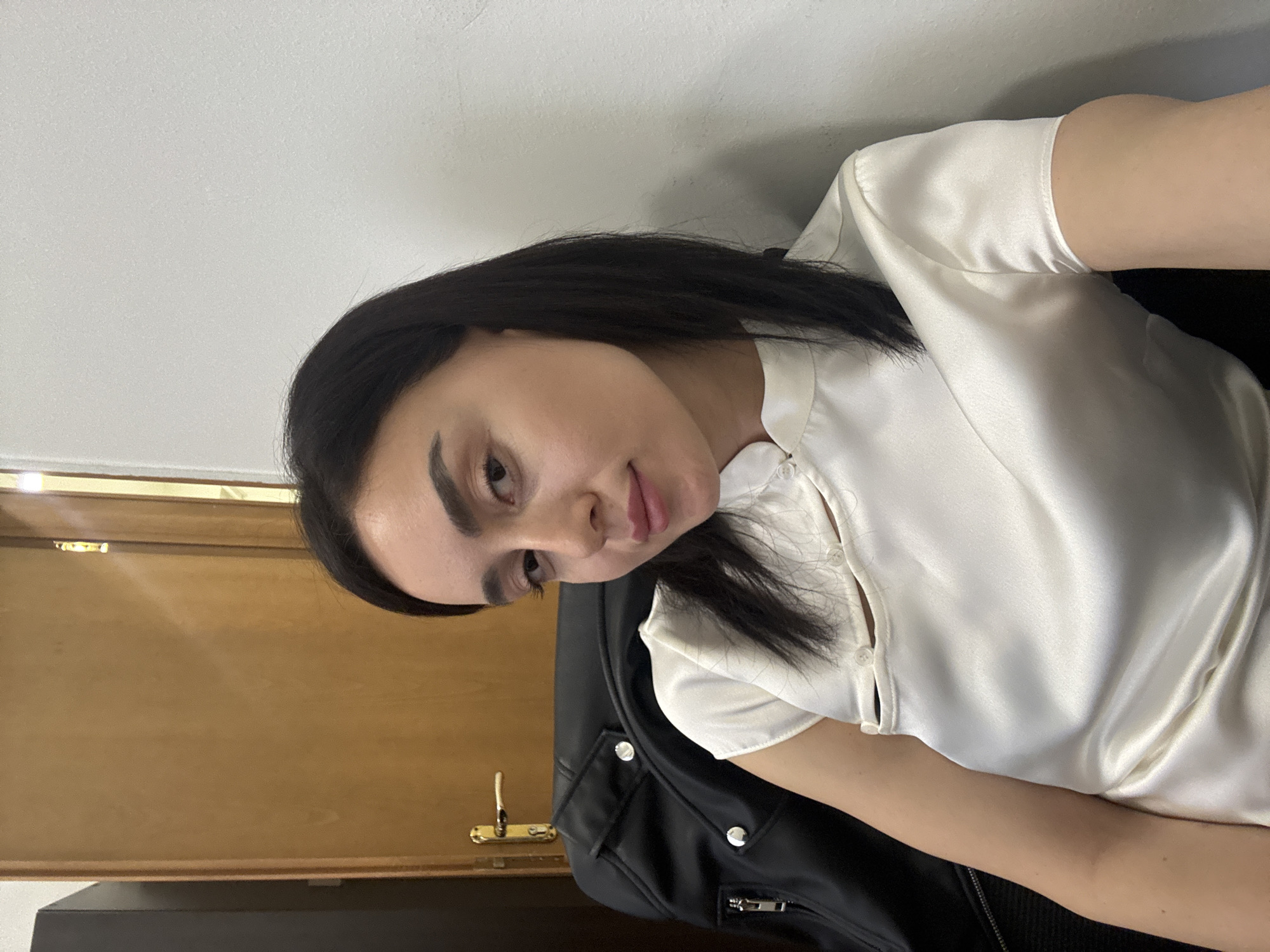 The image is a slightly angled, close-up shot of a woman indoors. She has dark hair and is wearing a white blouse. She appears to be sitting on or near a black leather jacket. Behind her, there is a wooden door with a gold-colored handle. The wall behind the door is white.
There is no information to determine if this picture was taken in the city of Tbilisi, Georgia.