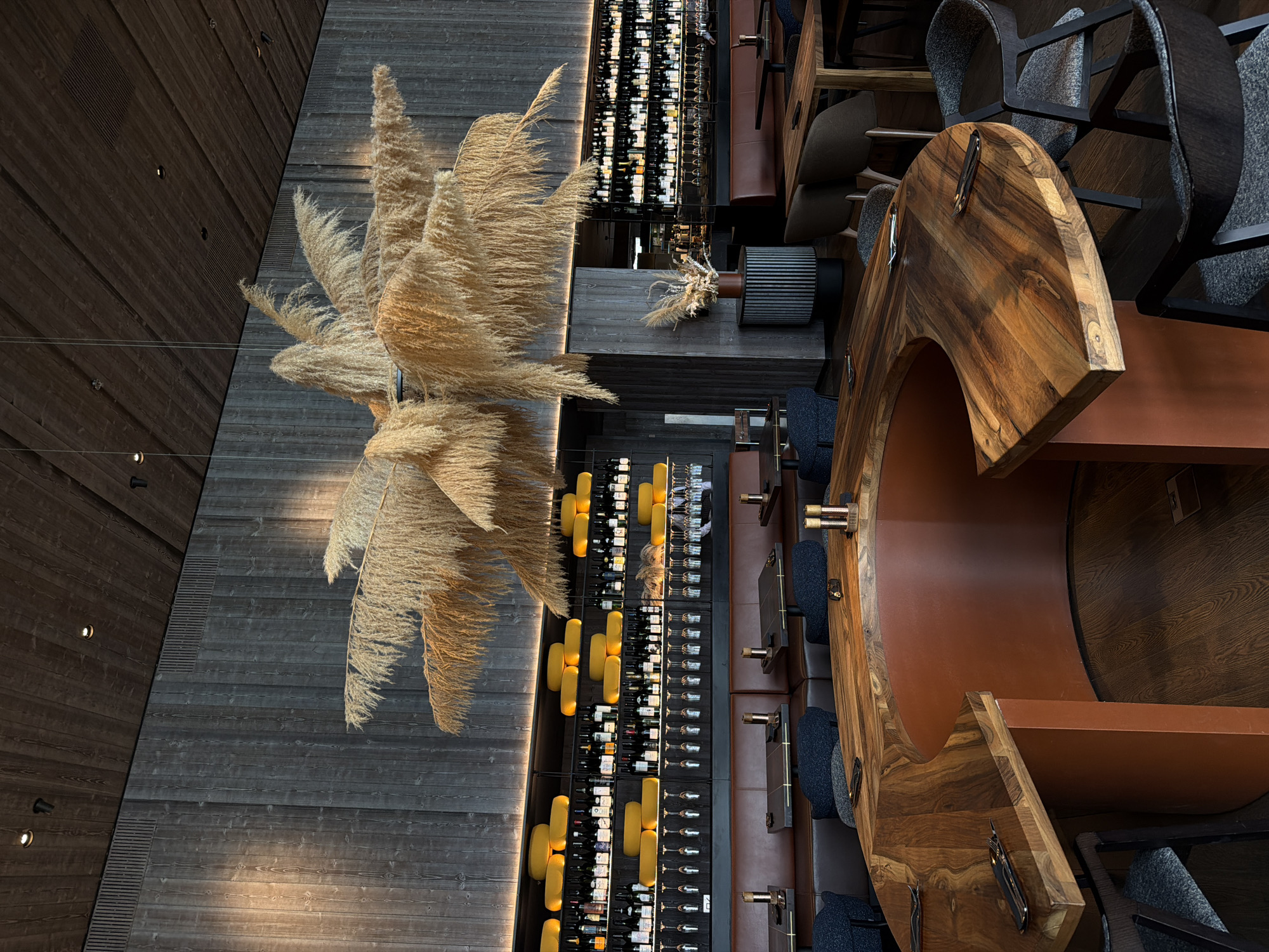 This image captures an overhead view of a sophisticated interior space, likely a bar or restaurant, characterized by a blend of rustic and modern design elements.
The central subject is a very large, voluminous decorative arrangement of dried, feathery pampas grass, suspended from the ceiling. Its light, golden-brown plumes cascade downwards, creating a striking visual focal point.
Below the pampas grass, a long, irregularly shaped bar counter crafted from dark, richly grained wood dominates the right side of the frame. Its unique, curved design, possibly resembling a hollowed-out log or a boat hull, is complemented by a lower, reddish-brown surface that forms the interior of the bar. Several bar stools with dark grey, textured fabric seats and black metal frames are positioned along the counter.
To the left of the bar, running vertically along a dark wooden wall, is an extensive wine display. Hundreds of wine bottles are arranged neatly on illuminated racks. Interspersed within the wine display are distinct yellow, cylindrical lighting fixtures that provide warm illumination. Adjacent to the wine bottles, another section displays rows of various glasses. Further down, additional seating areas with dark brown upholstered booths are visible.
The walls and ceiling are paneled with horizontal wooden planks, varying in tone from dark brown to lighter grey, adding depth and texture to the space. Small, recessed spotlights are integrated into the wooden surfaces, casting warm glows and highlighting the architectural details.
No people are visible, suggesting the scene is set for potential patrons. The overall ambiance conveyed is one of refined elegance and warmth, ideal for an evening setting, given the intentional lighting design. There is no visible text in the image.