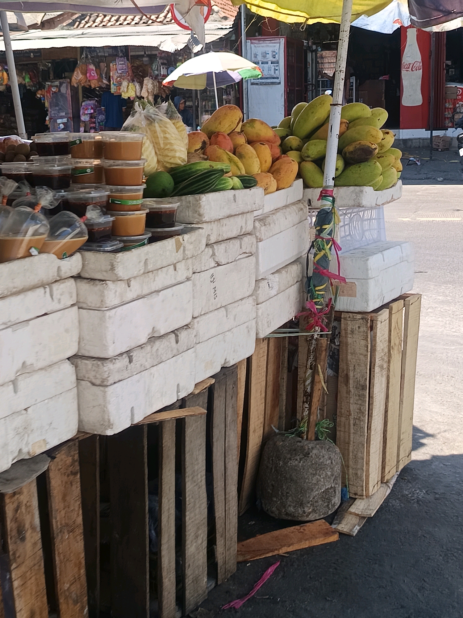 A roadside fruit stand in Blega, Indonesia, is depicted from a low angle, focusing on a display of produce. The stand is constructed from stacked white styrofoam blocks and wooden crates, supporting a variety of fruits and what appear to be preserved food items in plastic containers. Visible fruits include mangoes and what might be pineapples, along with green vegetables like cucumbers. A large, rough-hewn stone is placed at the base of the stand, possibly serving as a counterweight or decorative element. The background reveals a street scene with a building displaying a Coca-Cola logo, suggesting a commercial area. The lighting indicates it is daytime.