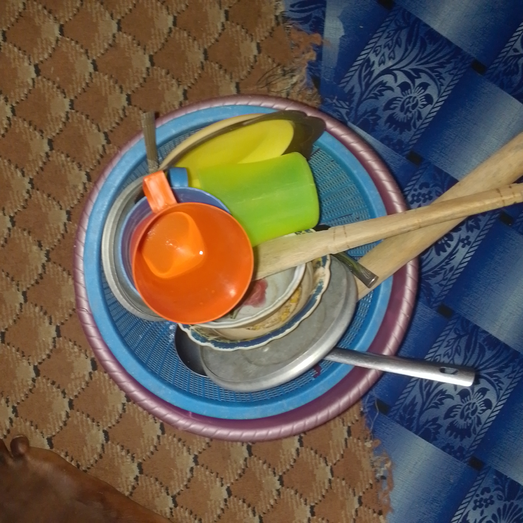 The image shows a top-down view of a blue plastic basket containing kitchen utensils. The basket is filled with several items, including an orange plastic ladle, a green plastic cup, and two wooden spoons or handles. Beneath these items, there are several metal bowls and plates, some of which appear to be stacked. The basket itself is placed on a patterned brown rug, with a blue fabric with a floral pattern visible to the right. The lighting suggests an indoor environment, likely a home. No specific details about the city of Jalingo, Nigeria are visible in the image.