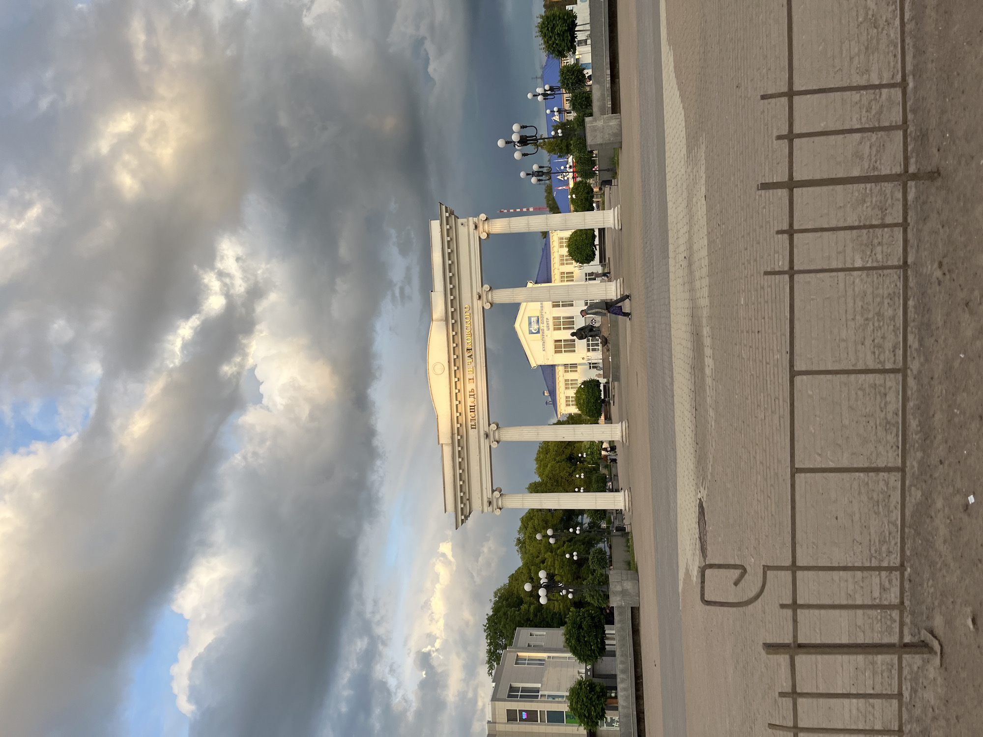 The image shows a street scene in Chaikovsky, Russia, with a white classical-style archway dominating the center. The archway has columns and inscription in Russian. Behind the archway is a white building with a blue roof. Two people are walking near the base of the archway. Streetlights are positioned along the street. The sky is filled with dark, dramatic storm clouds on one side and lighter, illuminated clouds on the other side. The foreground is a gray brick pavement with some markings, likely designating parking spaces. Other buildings and trees are visible in the background.