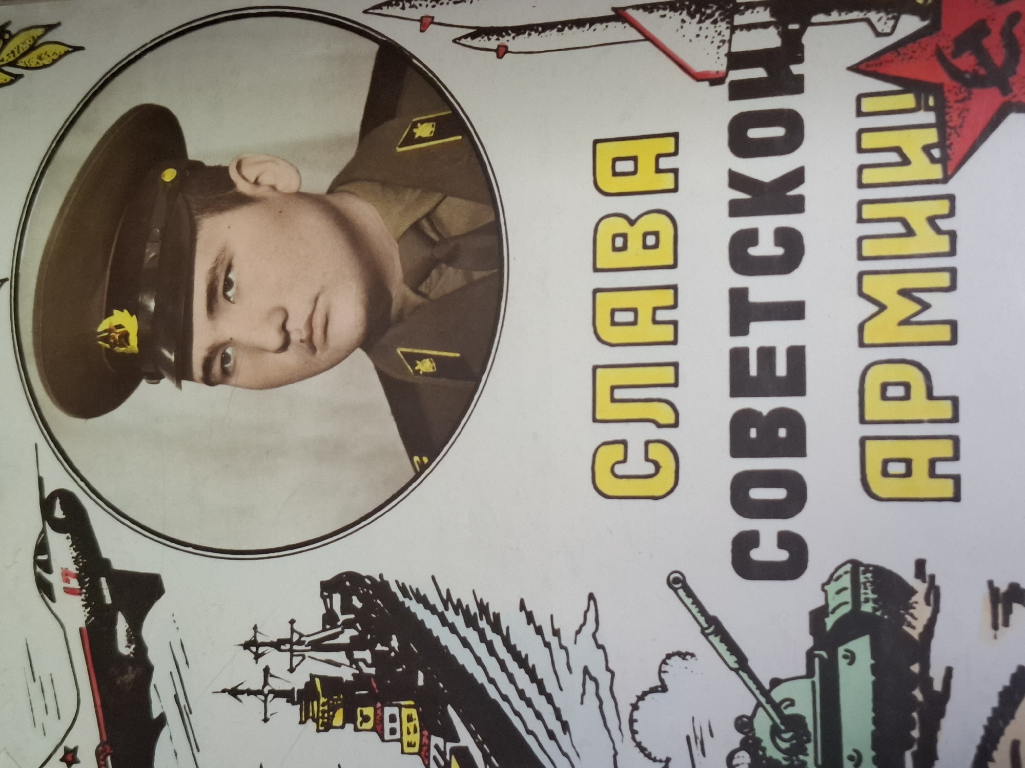 The image is a close-up of a Soviet-era propaganda poster. The poster features a portrait of a young man in a military uniform and cap, set within a circular frame. To the left of the portrait are illustrations of military hardware, including a fighter jet in the upper left and a warship below it. To the right of the portrait, there are stylized depictions of rockets or missiles. In the foreground, towards the bottom left, is an illustration of a tank.
Below the portrait, large text in Cyrillic characters reads "СЛАВА СОВЕТСКОЙ АРМИИ!", which translates to "Glory to the Soviet Army!". This text is yellow with a dark outline. A red star with a hammer and sickle emblem is visible in the lower right corner.
The poster has a white background with some visible texture and possibly some minor blemishes. The lighting appears to be from a natural source, casting soft shadows.
There is no discernible information in the image that indicates its location within the city of Almaty, Kazakhstan. The content is purely political and military propaganda, typical of the Soviet Union.